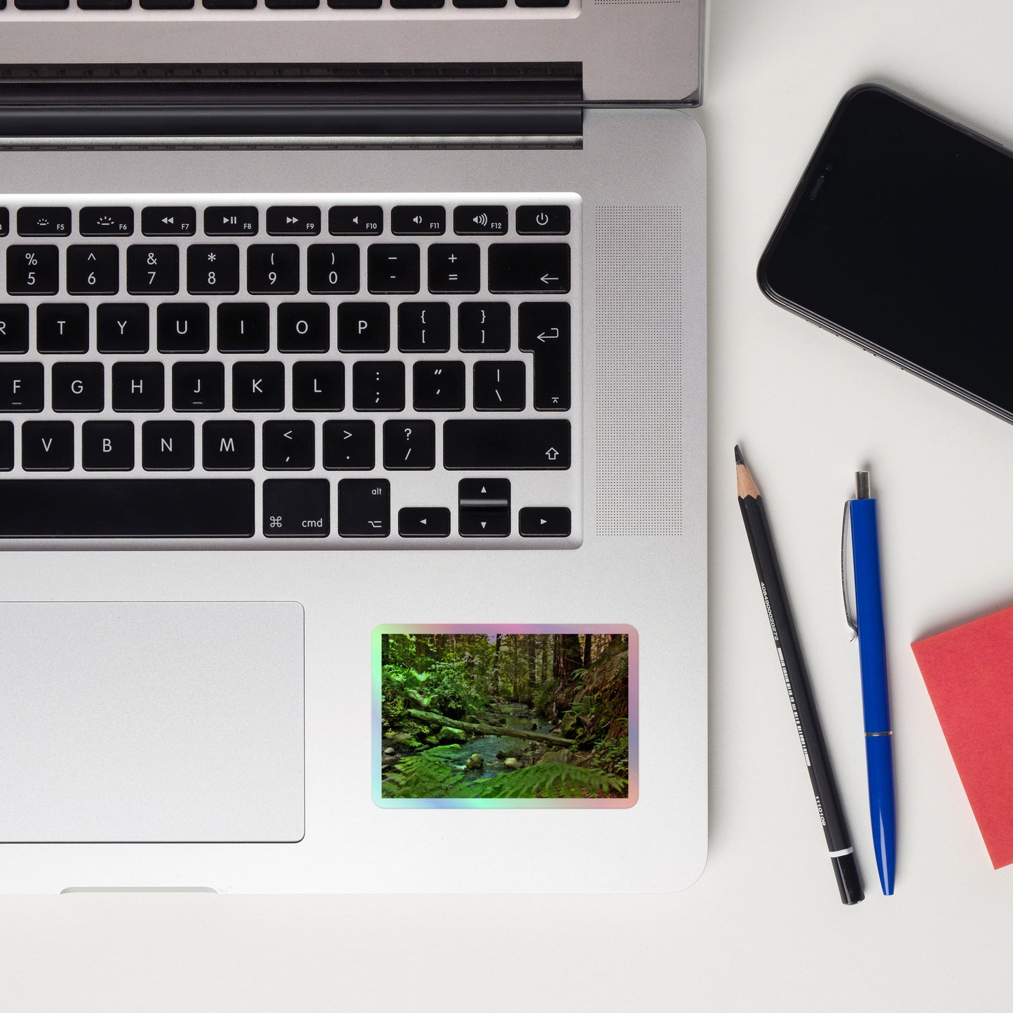 Lifestyle photo of a rectangular holographic sticker with an image of a creek through a redwood forest with ferns, placed on a laptop with desk supplies nearby.