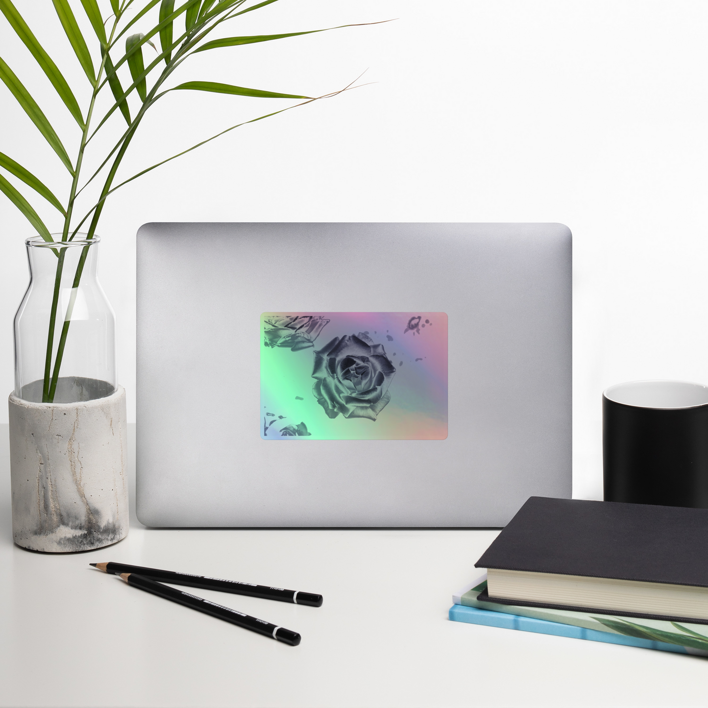 Lifestyle photo of a rectangular holographic sticker with an image of a silver reflective rose design, placed on a laptop with desk supplies nearby.