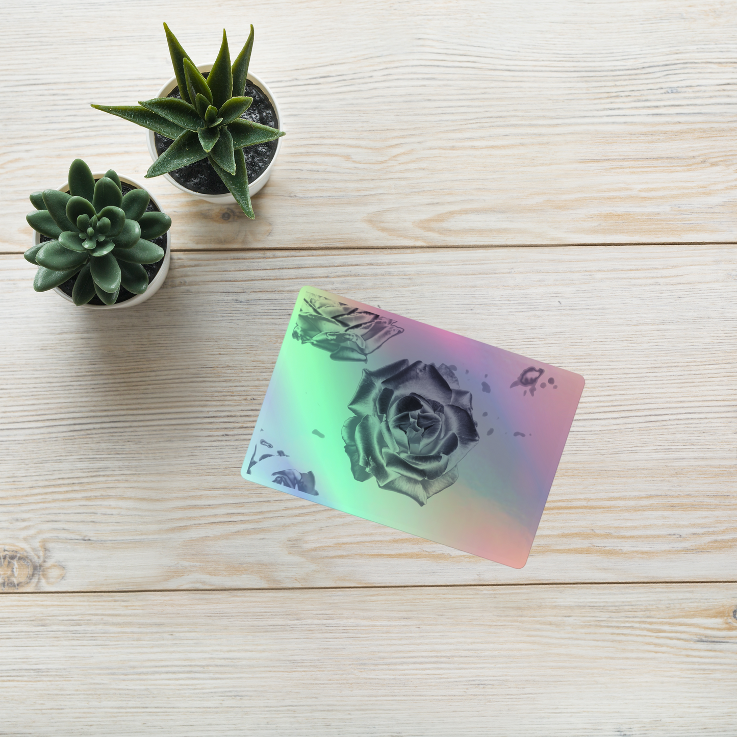 Lifestyle photo of a rectangular holographic sticker with an image of a silver reflective rose design, placed on a table with two small succulents.