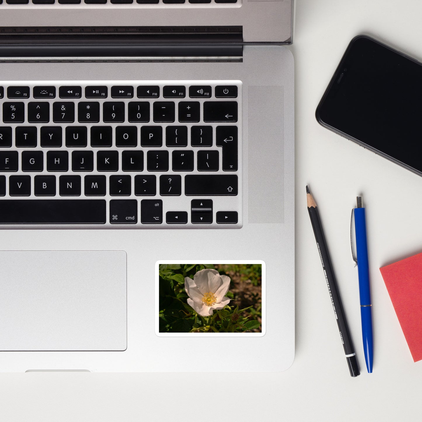 Lifestyle photo of a rectangular vinyl sticker with an image of white rose bloom with yellow stamen, placed on a laptop to the right of the mouse pad.