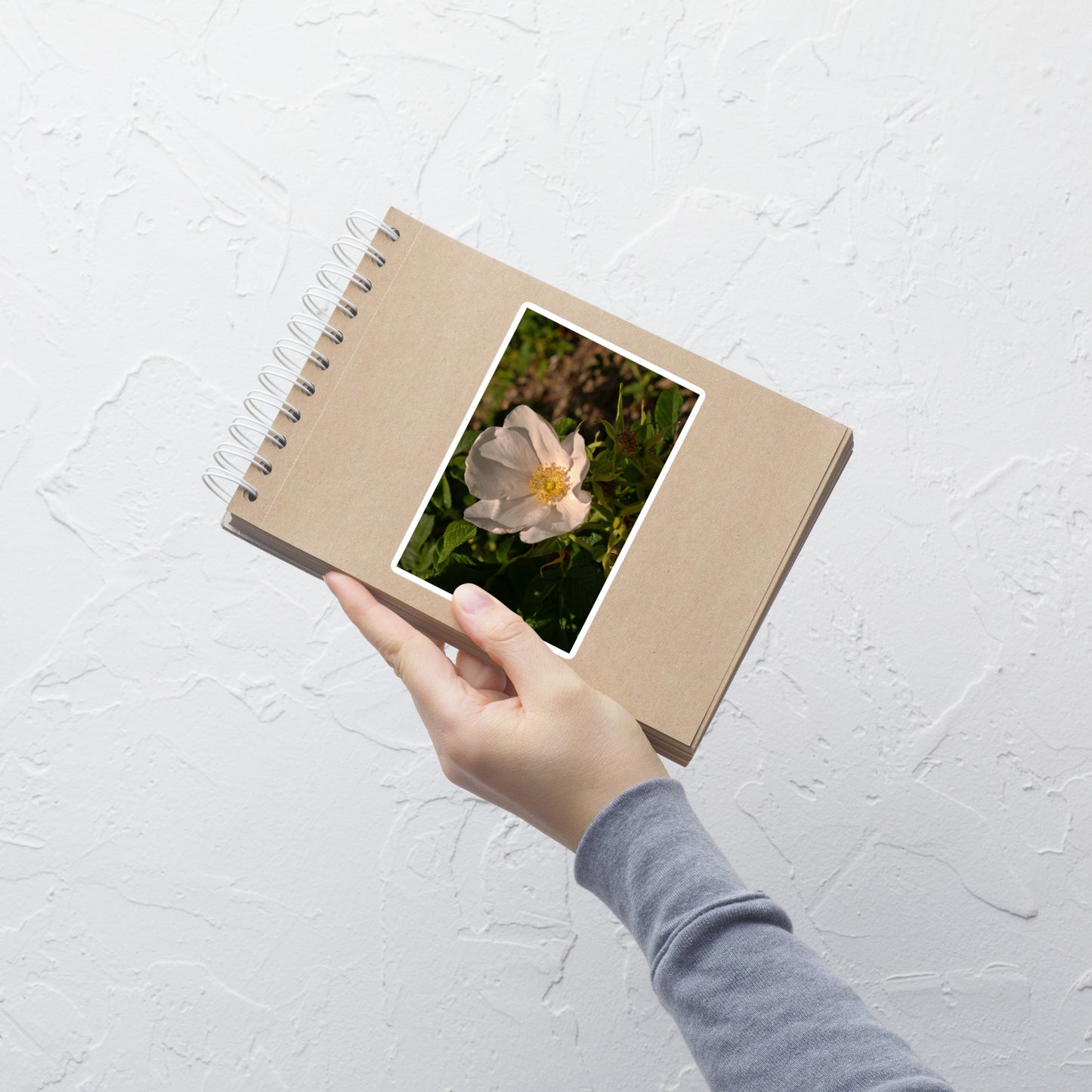Lifestyle photo of a rectangular vinyl sticker with an image of white rose bloom with yellow stamen, placed on a small notepad.