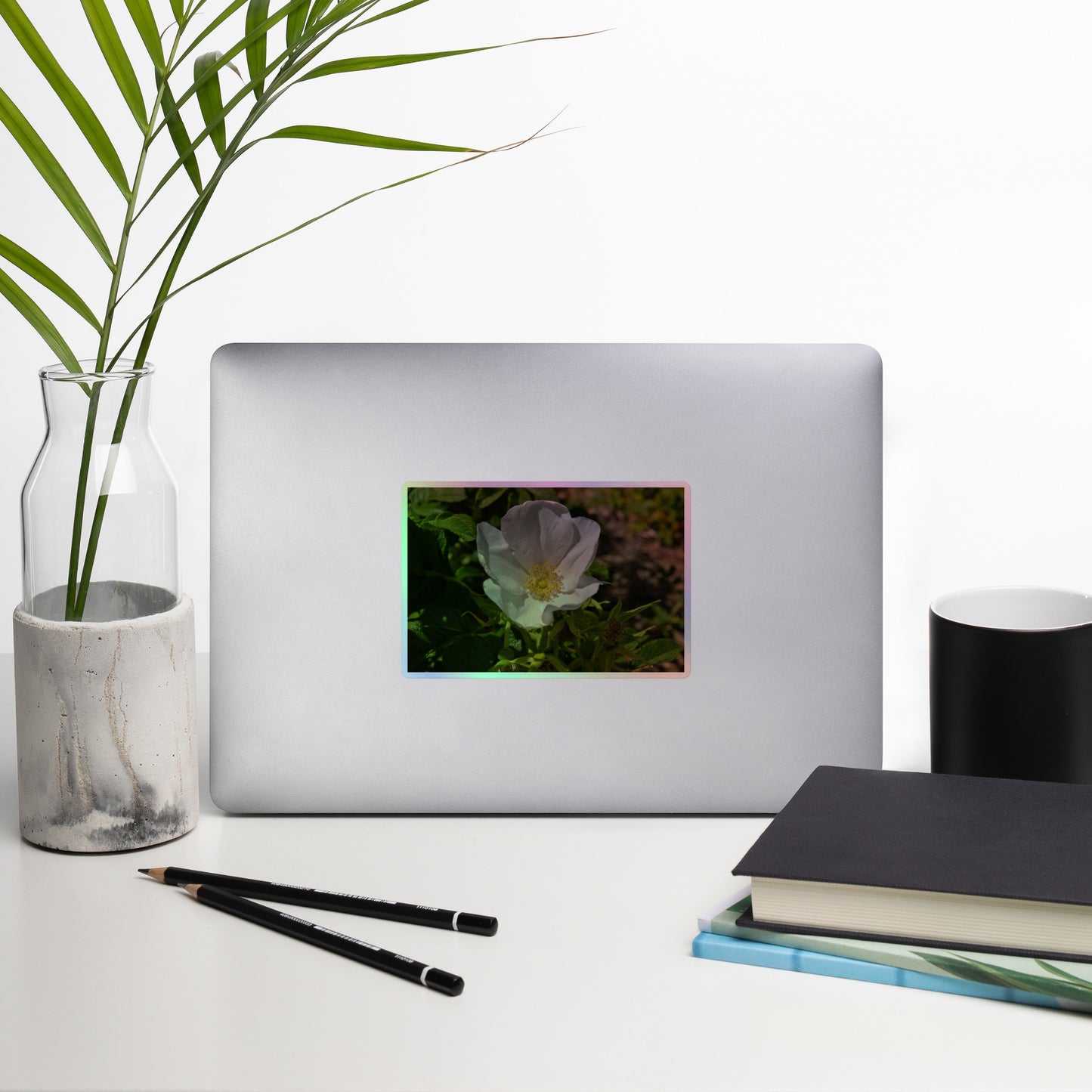 Lifestyle photo of a rectangular holographic sticker with a close-up image of a white flower with yellow stamen, placed on a laptop with desk supplies nearby.