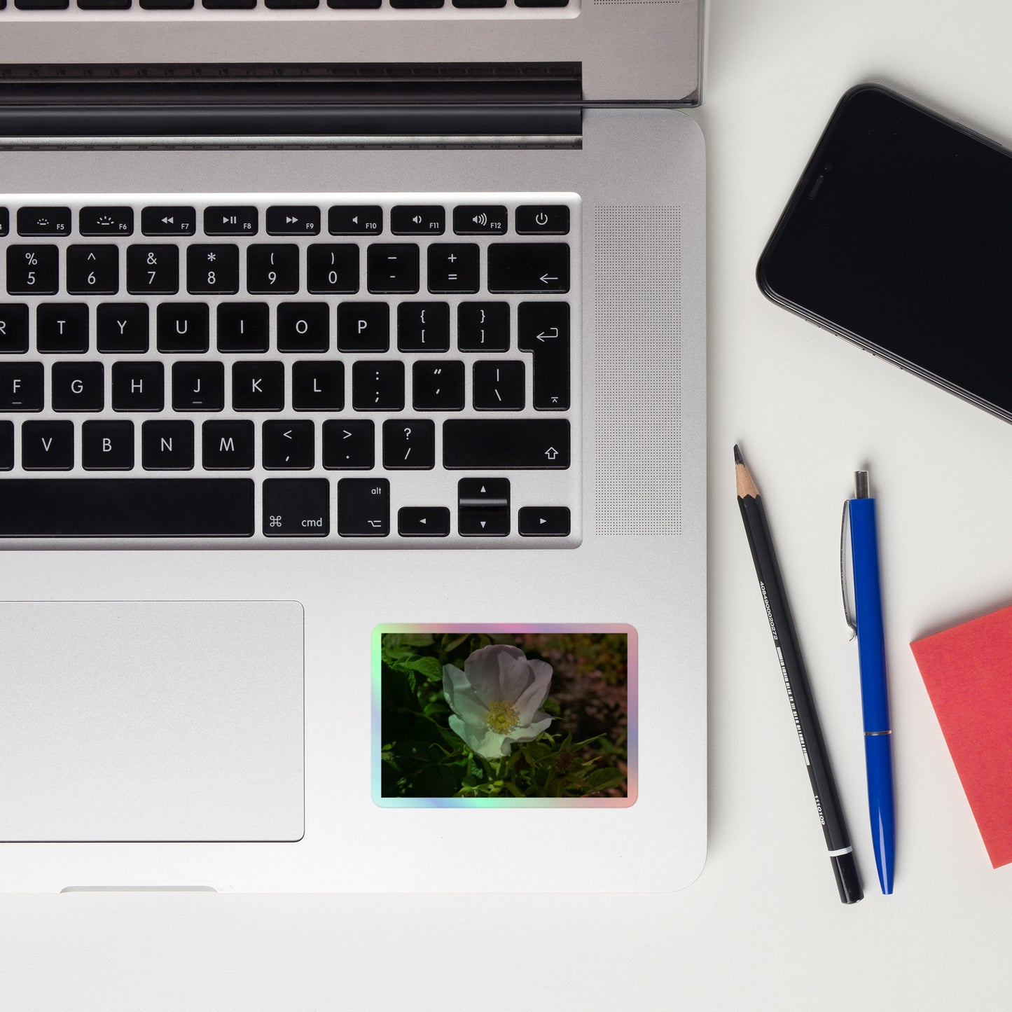 Lifestyle photo of a rectangular holographic sticker with a close-up image of a white flower with yellow stamen, placed on a laptop to the right of the mouse pad.