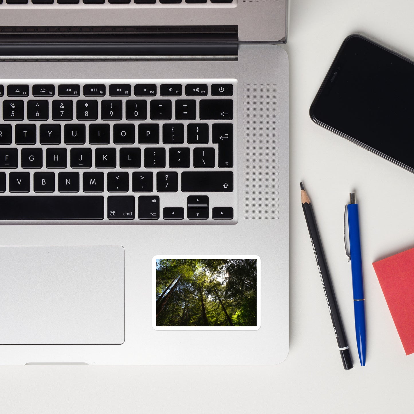 Lifestyle photo of a rectangular vinyl sticker with an image of a circle of redwood trees as seen from below, placed on a laptop to the right of the mouse pad.