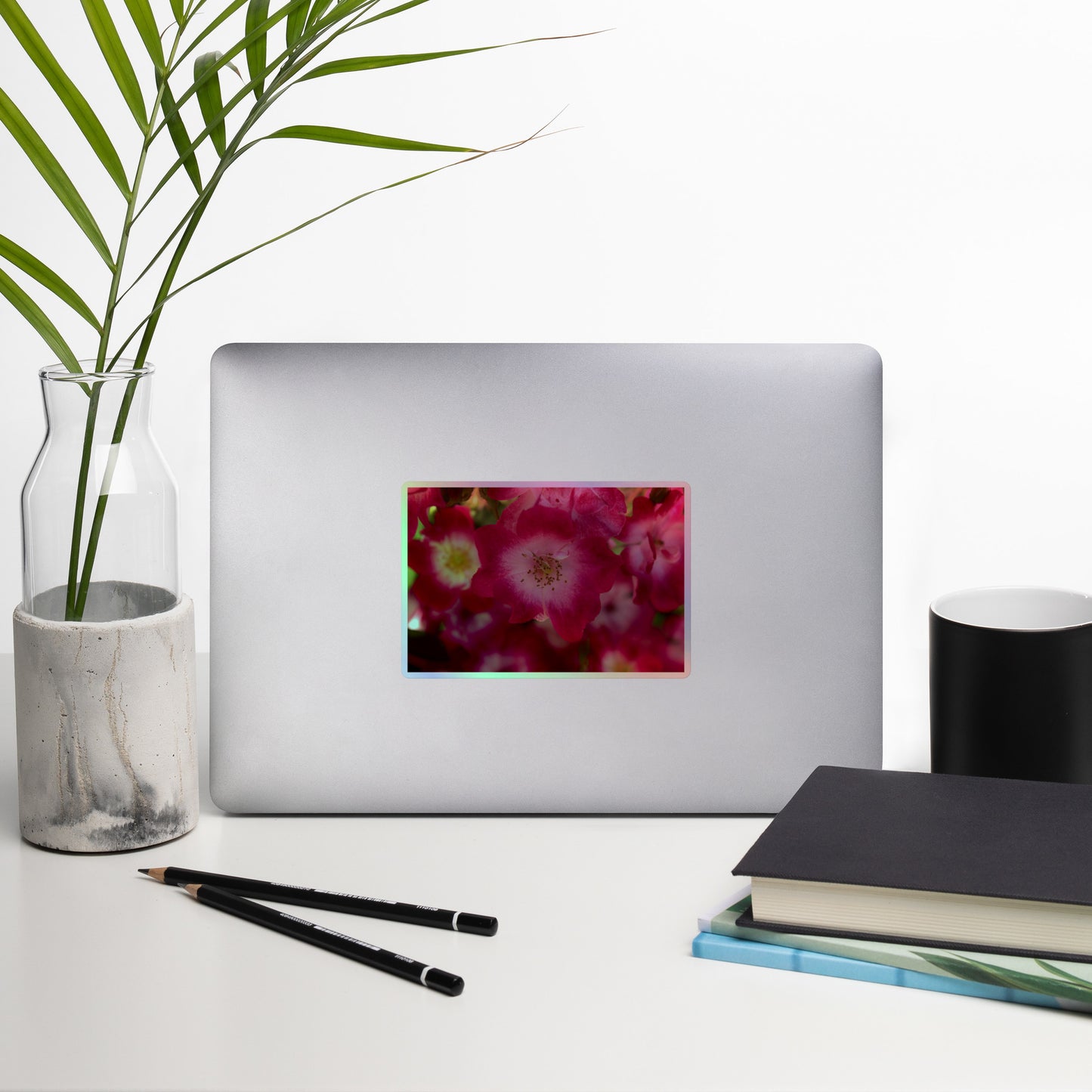 Lifestyle photo of a rectangular holographic sticker with an image of pink rose blooms with white centers, placed on a laptop with desk supplies nearby.
