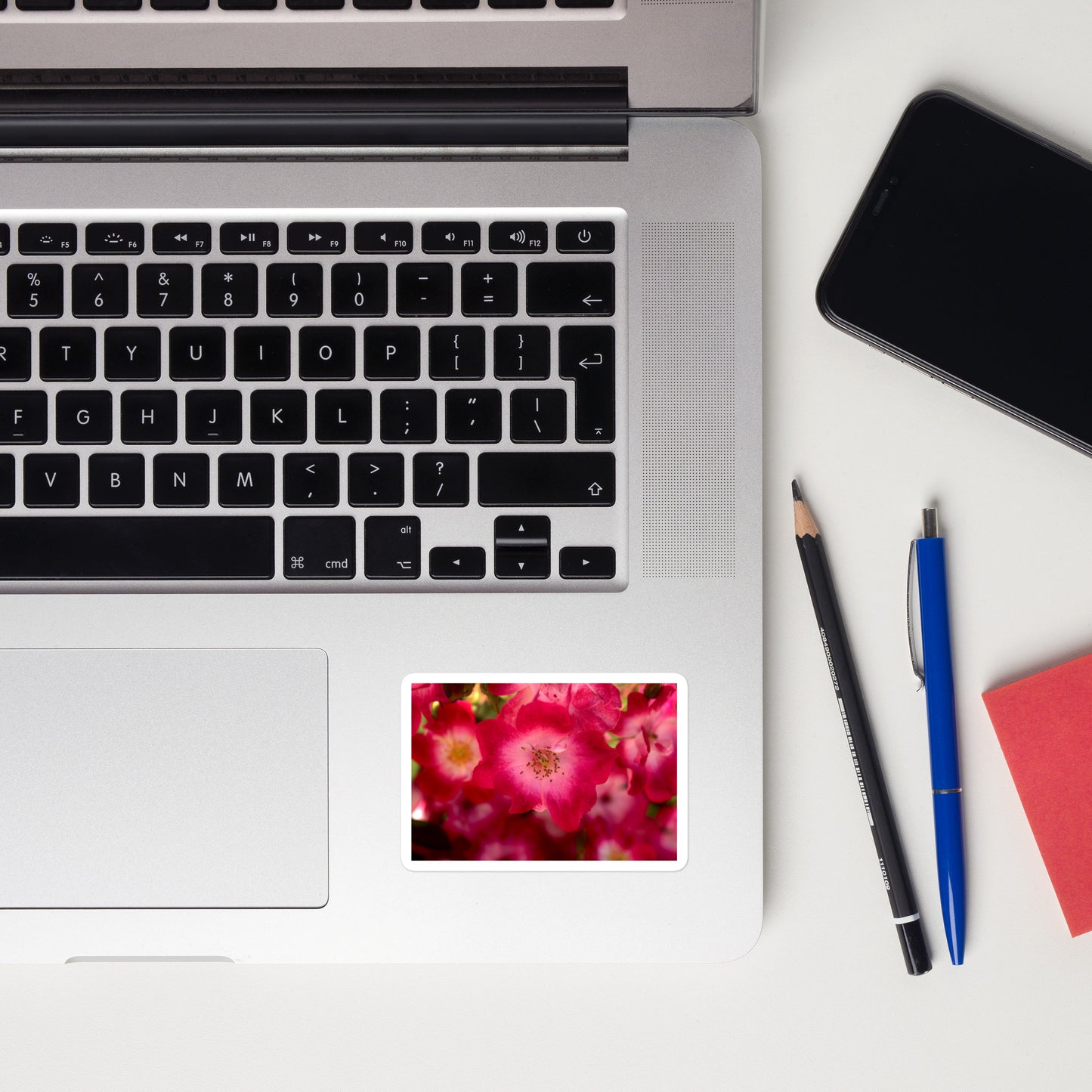 Lifestyle photo of a rectangular vinyl sticker with an image of dark pink flowers with white centers, placed on a laptop to the right of the mouse pad.