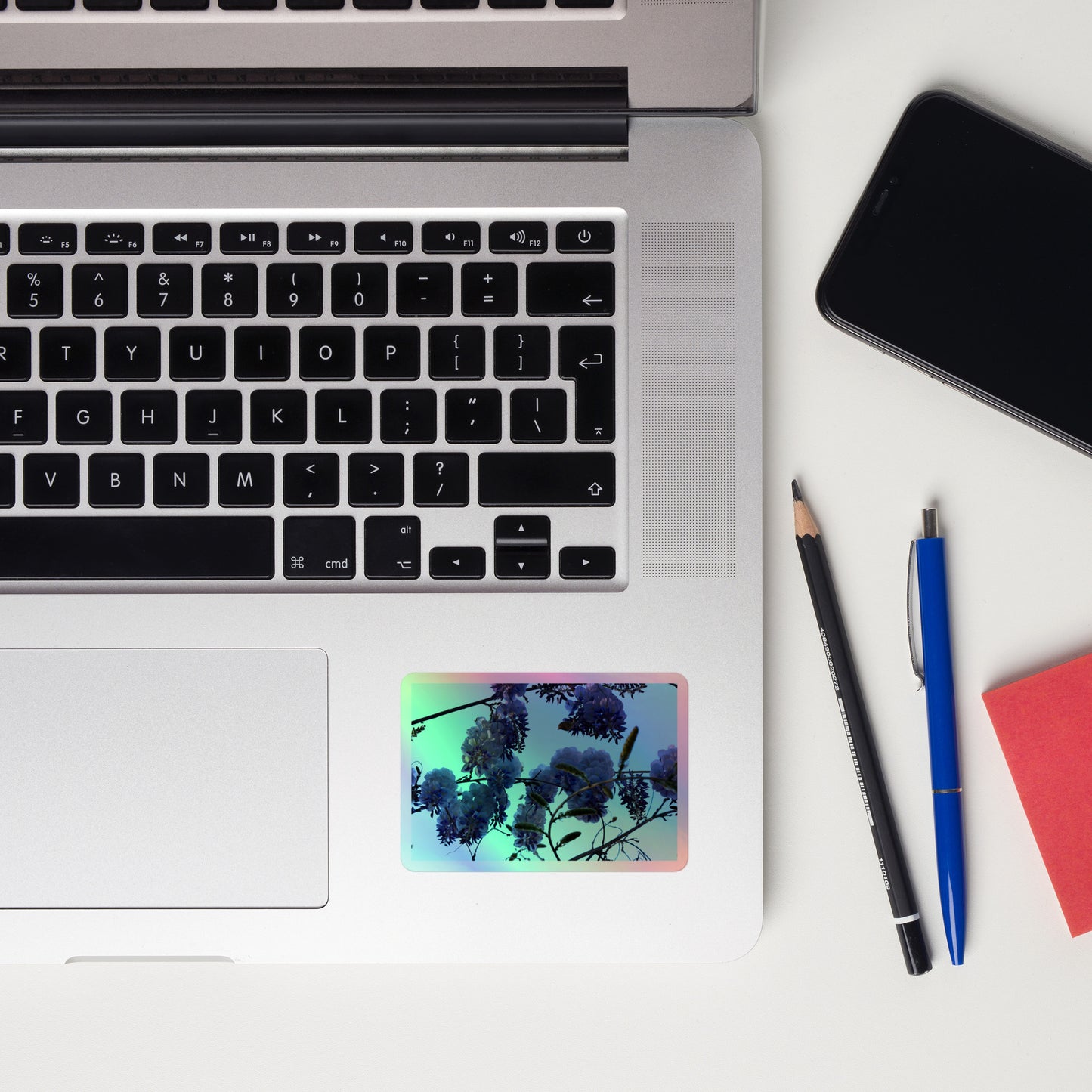 Lifestyle photo of a rectangular holographic sticker with an image of purple wisteria blooms with blue sky in the background, placed on a laptop to the right of the mouse pad.