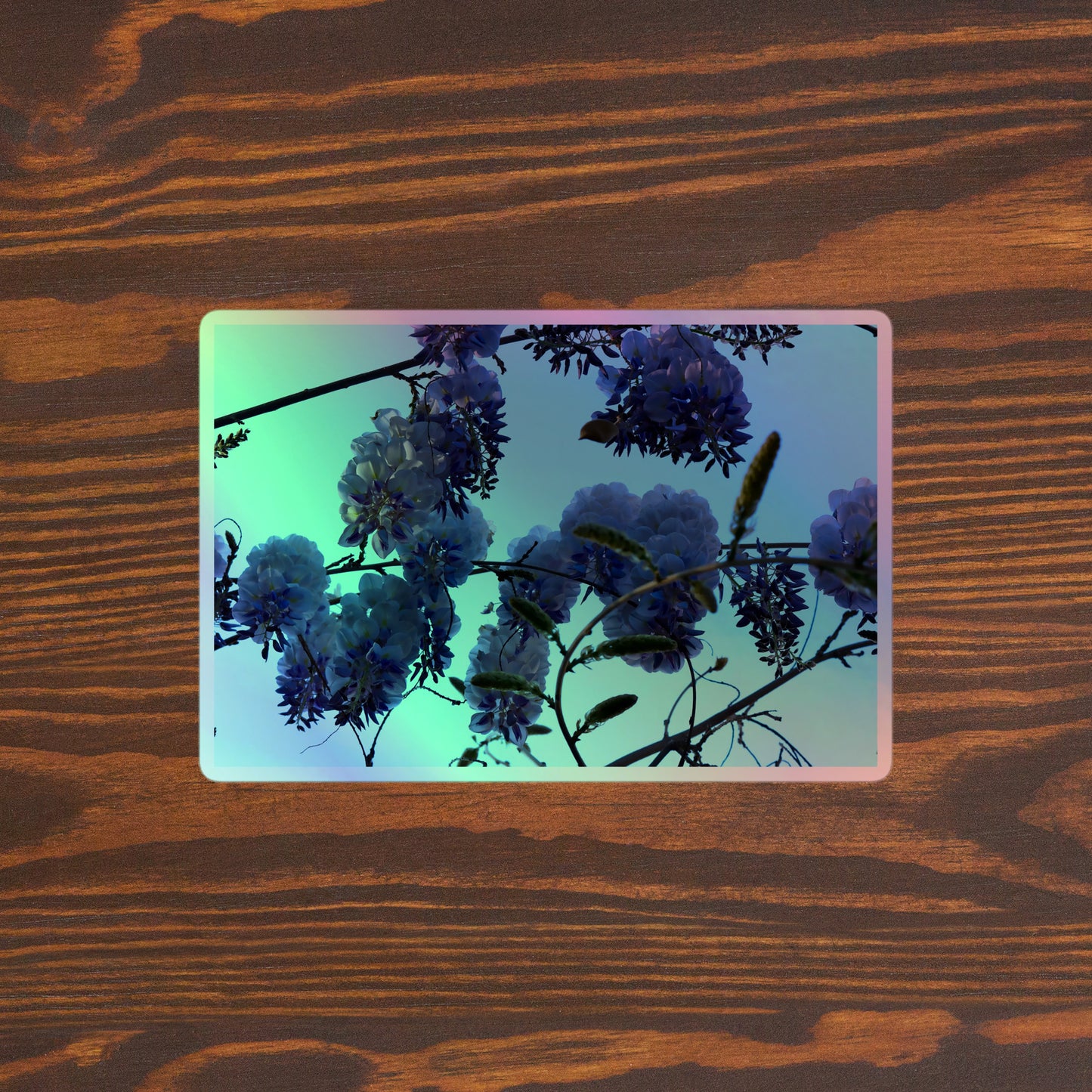 Lifestyle photo of a rectangular holographic sticker with an image of purple wisteria blooms with blue sky in the background, placed on a brown wooden table surface.