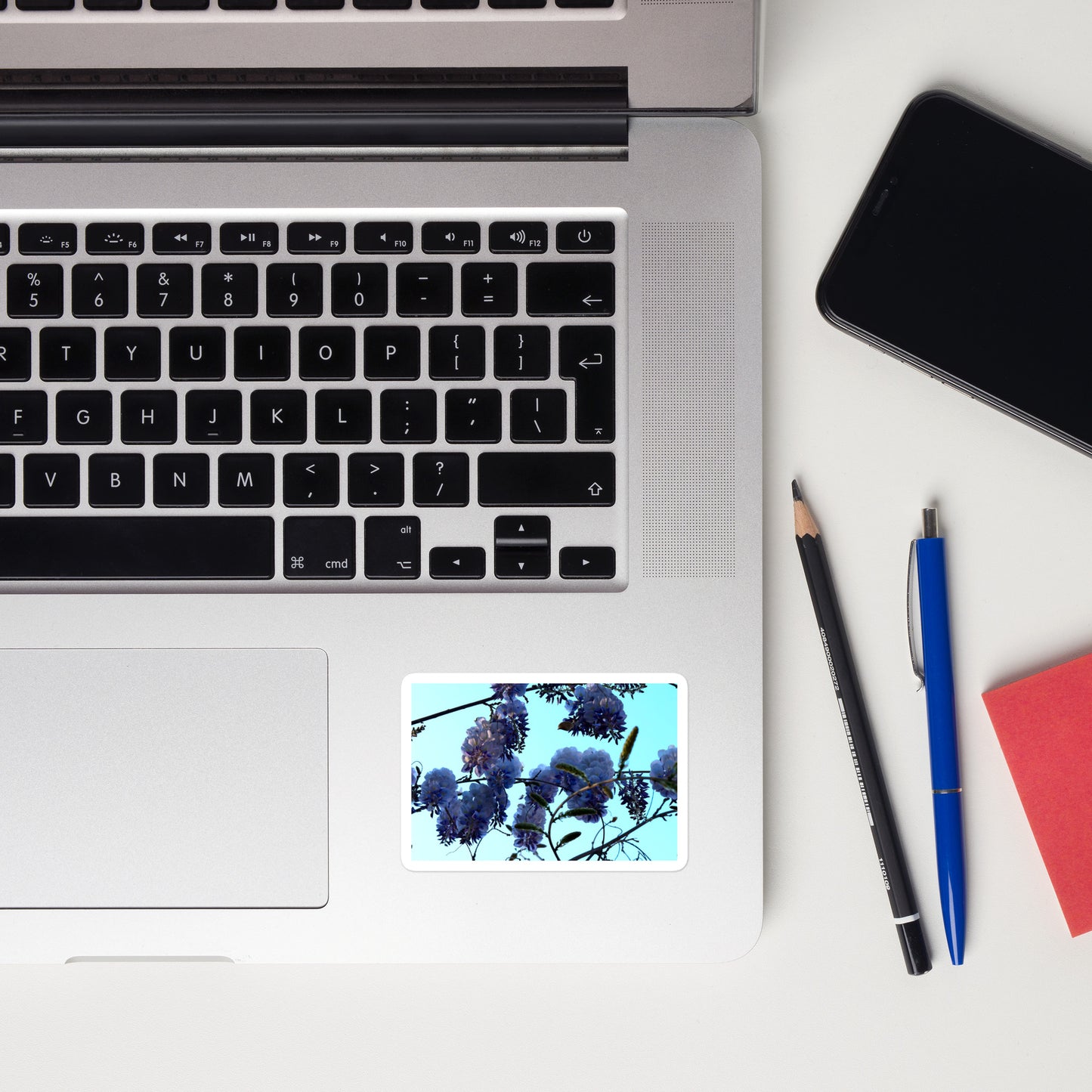 Lifestyle photo of a rectangular vinyl sticker with an image of a blue sky and purple wisteria blooms, placed on a laptop to the right of the mouse pad.
