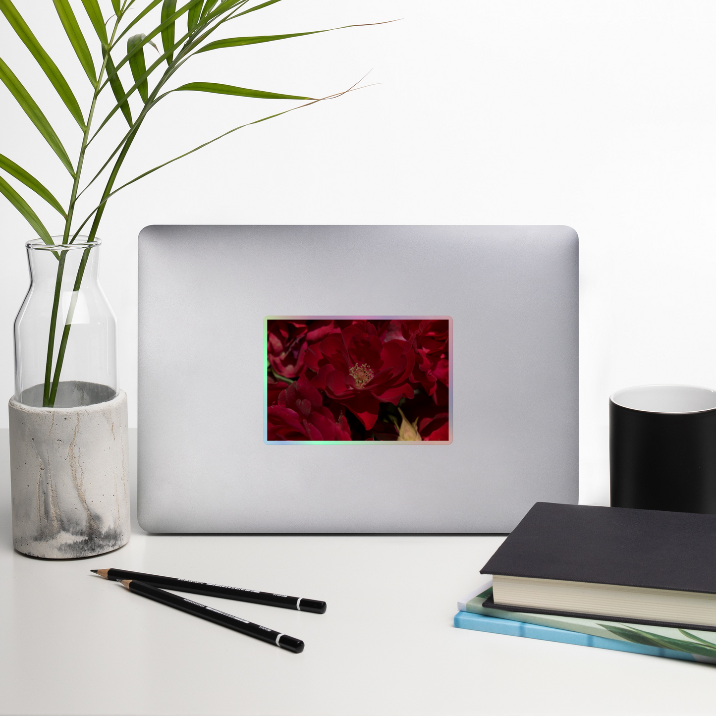 Lifestyle photo of a rectangular holographic sticker with a close-up image of dark red roses, placed on a laptop with desk supplies nearby.