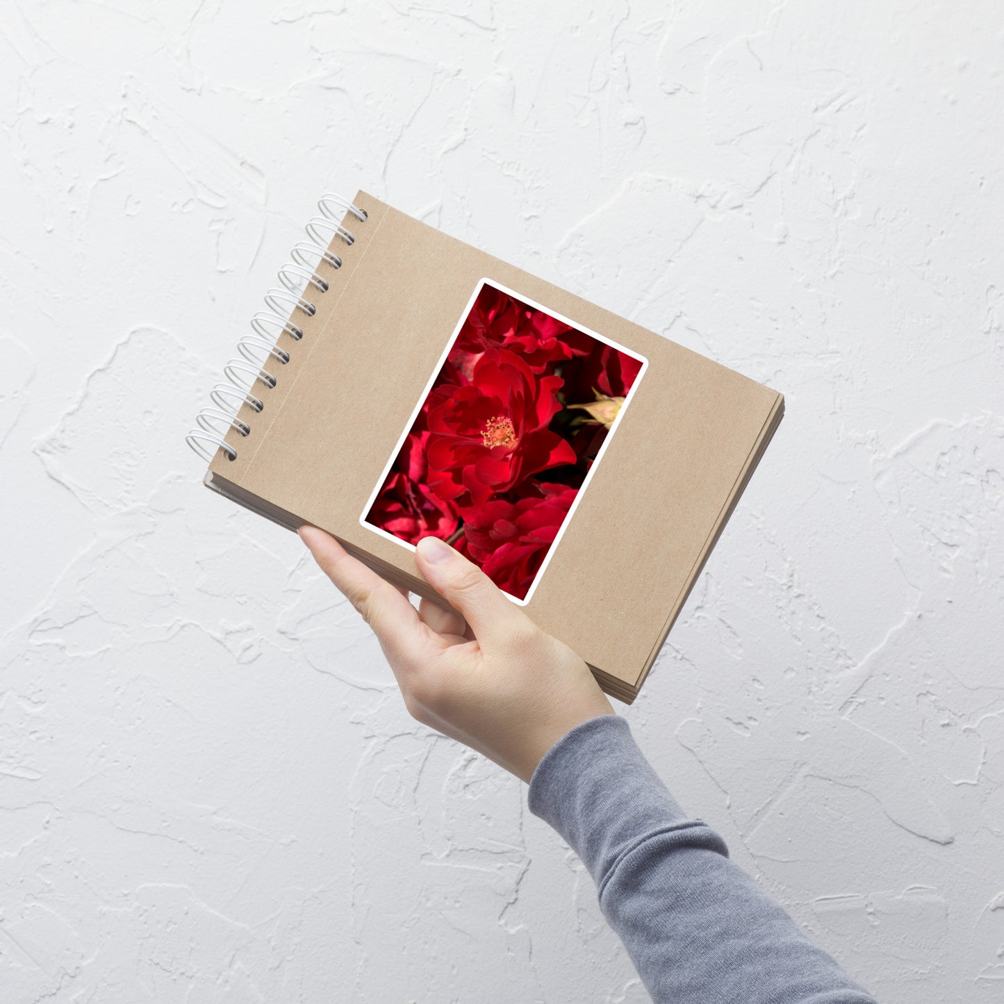 Lifestyle photo of a rectangular vinyl sticker with an image of bright red rose blooms with visible stamen, placed on a small notepad.