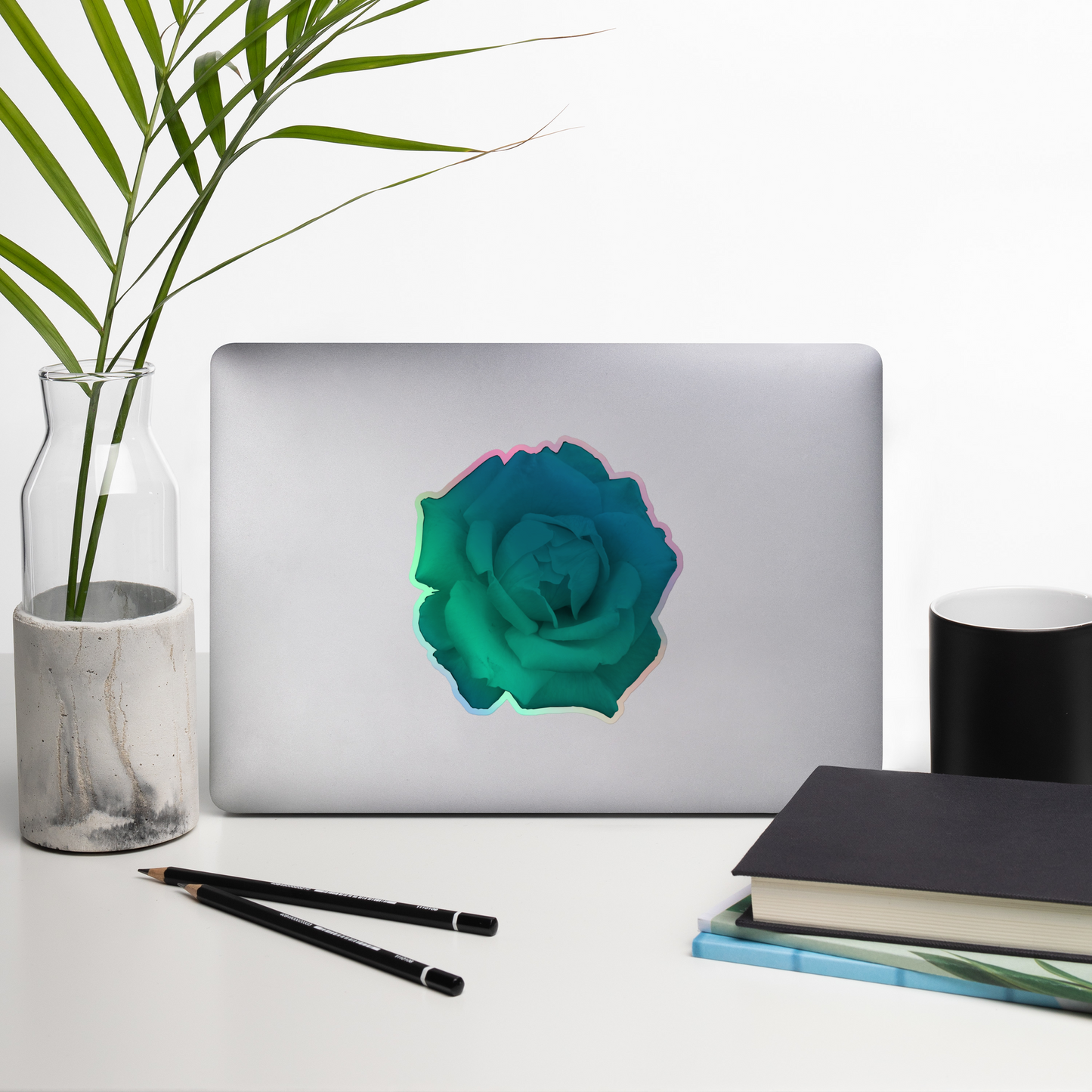 Lifestyle photo of a kiss-cut holographic sticker with an image of a vivid turquoise rose design, placed on a laptop with desk supplies nearby.