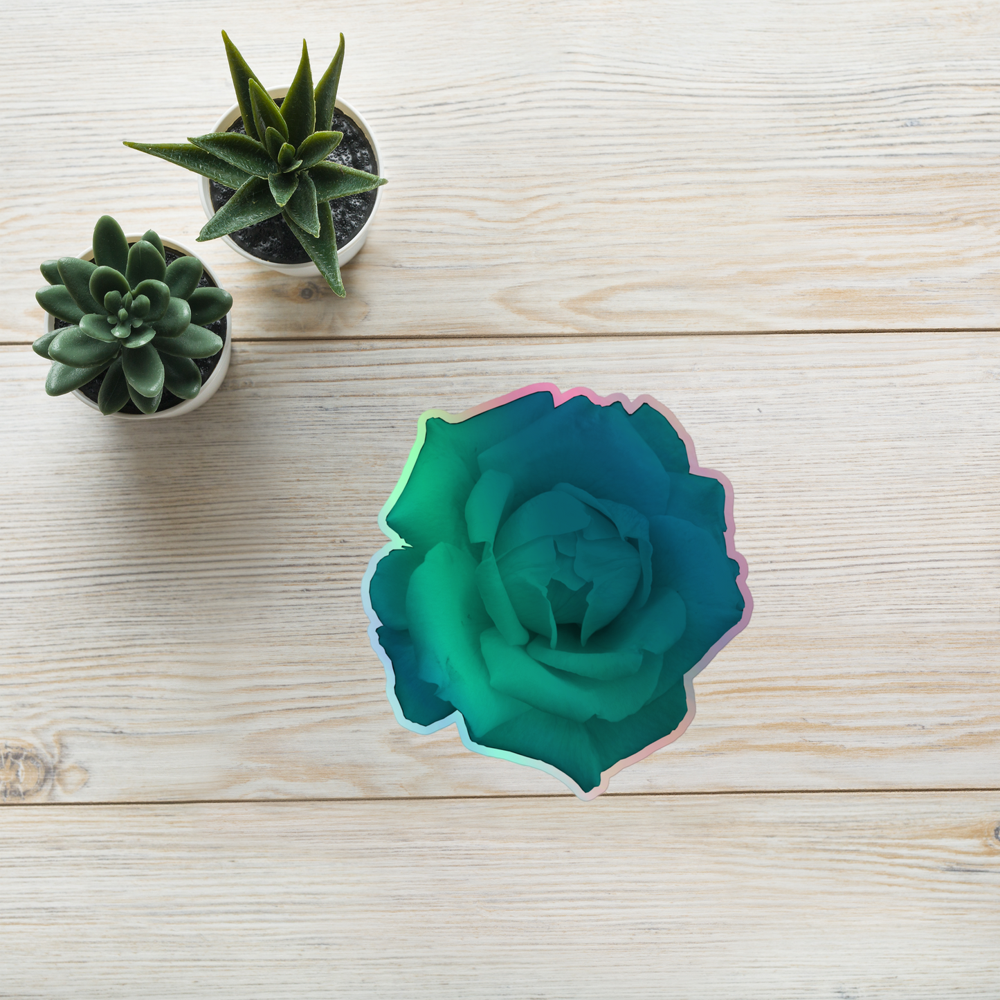 Lifestyle photo of a kiss-cut holographic sticker with an image of a vivid turquoise rose design, placed on a table with two small succulents.