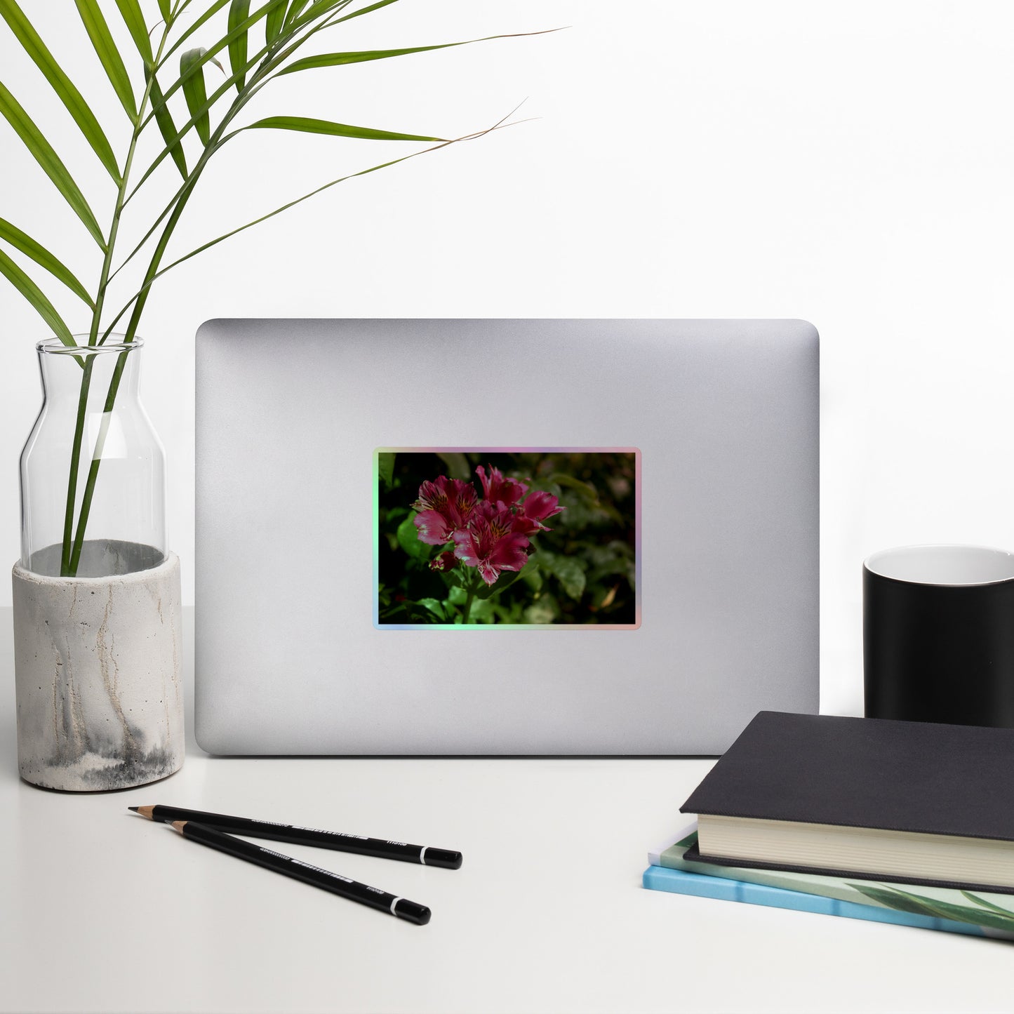 Lifestyle photo of a rectangular holographic sticker with a close-up image of vivid pink Peruvian Lilies with yellow accents, placed on a laptop with desk supplies nearby.