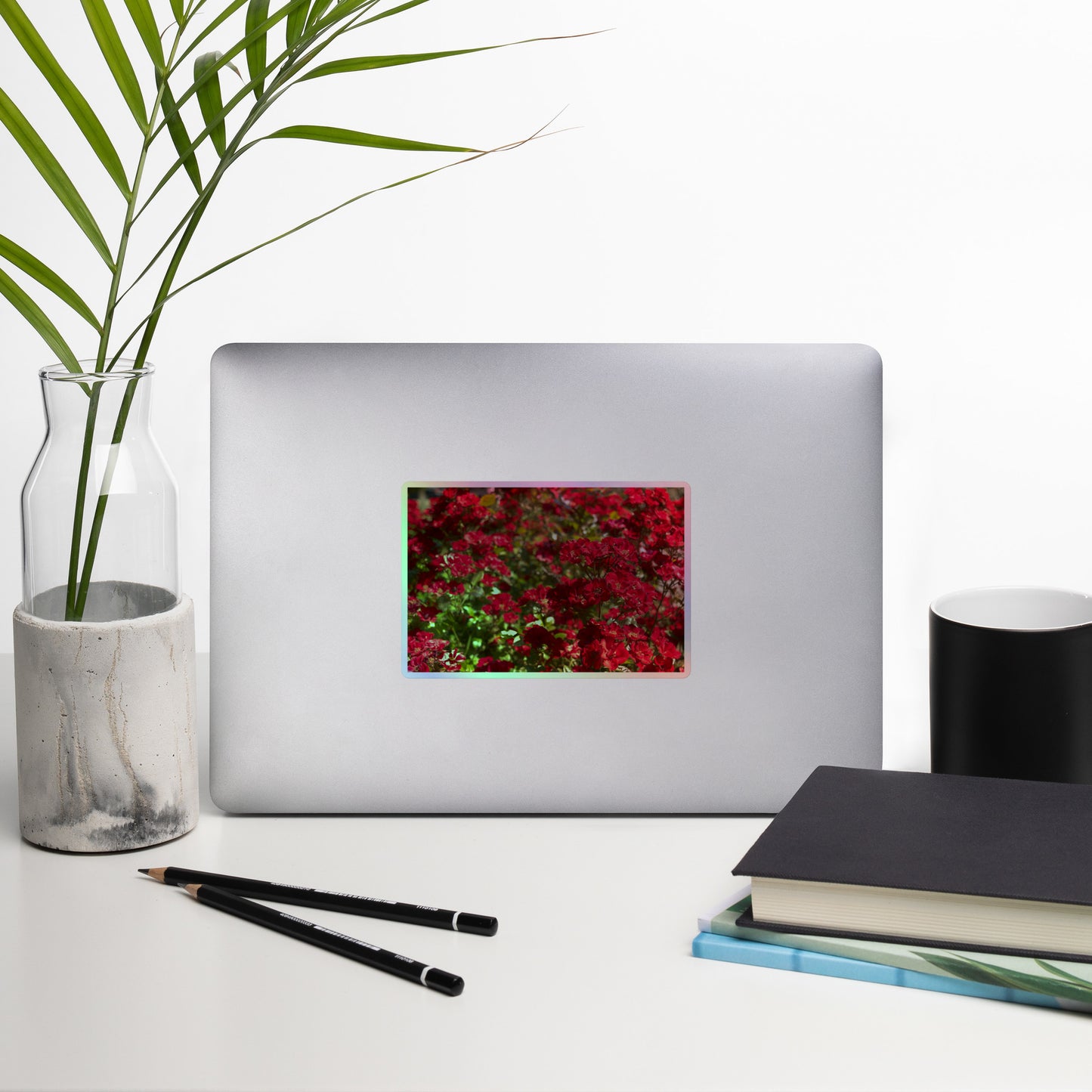 Lifestyle photo of a rectangular holographic sticker with an image of many small, bright red flowers, placed on a laptop with desk supplies nearby.