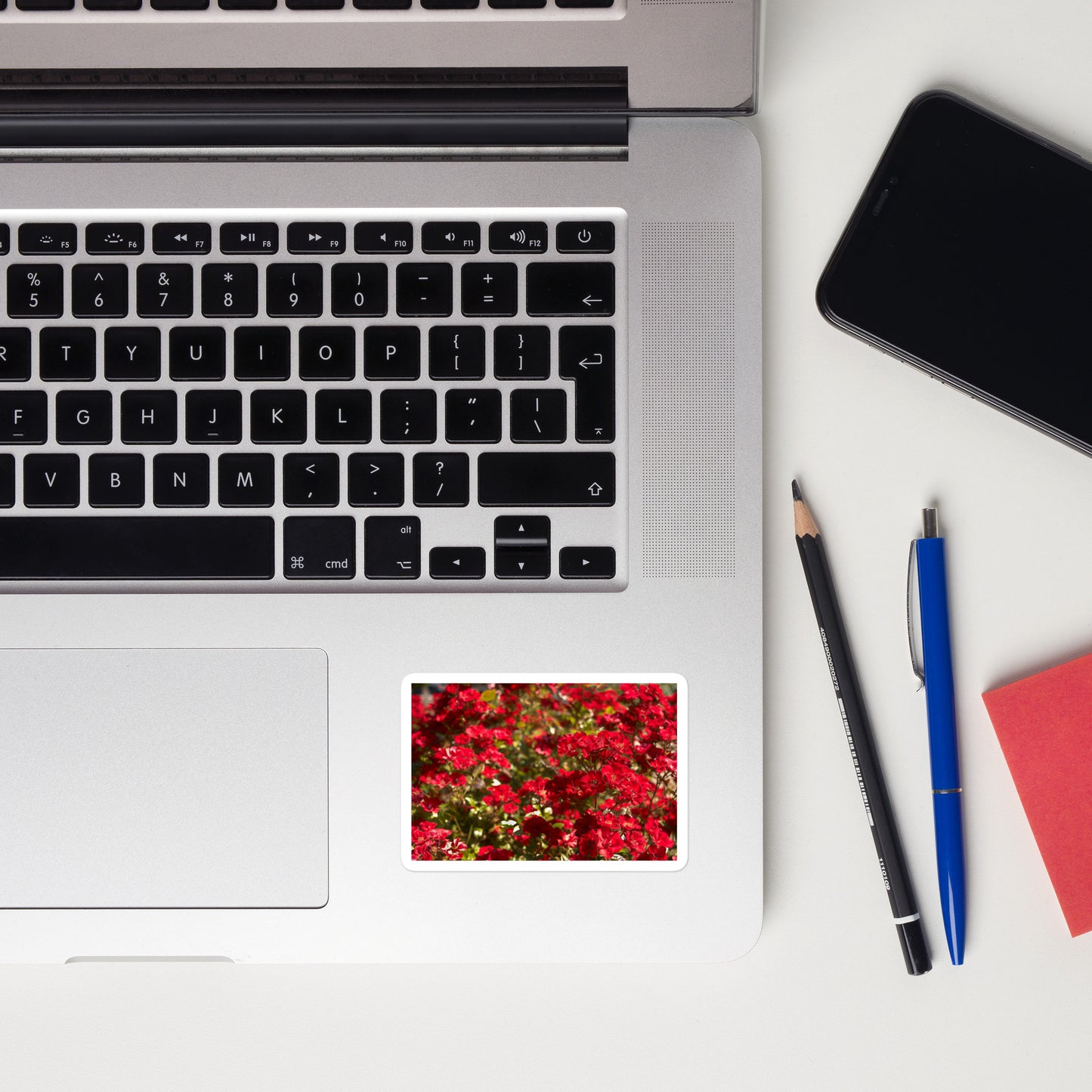 Lifestyle photo of a rectangular vinyl sticker with an image of a bush of bright red flowers, placed on a laptop to the right of the mouse pad.