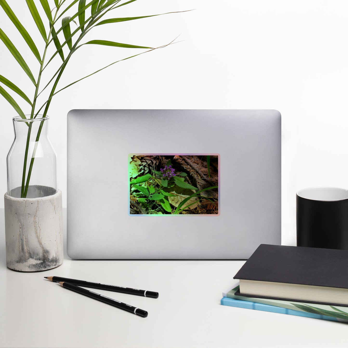 Lifestyle photo of a rectangular holographic sticker with a close-up image of a purple Heal-All flower in a redwood forest floor, placed on a laptop with desk supplies nearby.