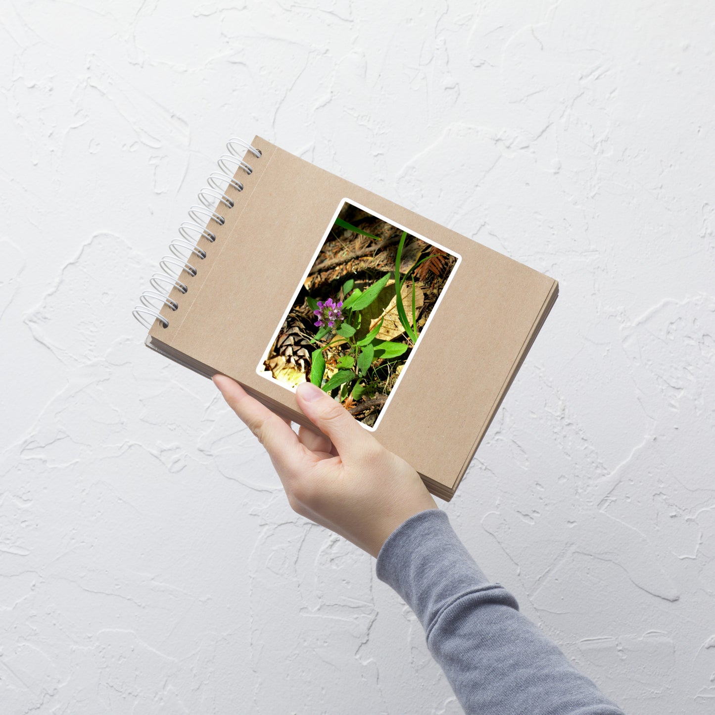 Lifestyle photo of a rectangular vinyl sticker with an image of a purple wild flower in a forest floor, placed on a small notepad.