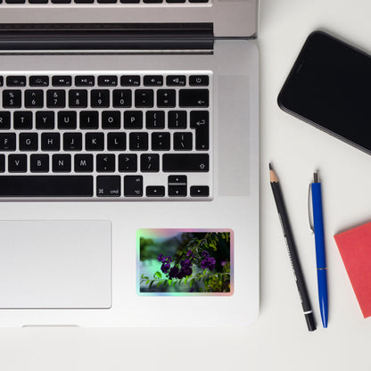 Lifestyle photo of a rectangular holographic sticker with an image of purple flowers on a tree branch, placed on a laptop to the right of the mouse pad.