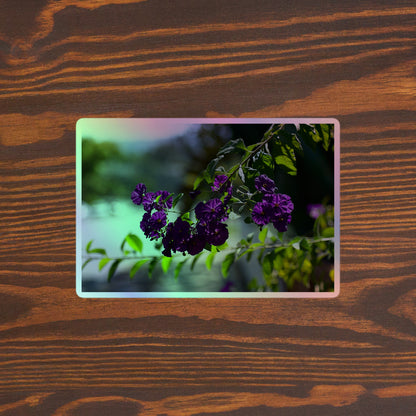 Lifestyle photo of a rectangular holographic sticker with an image of purple flowers on a tree branch, placed on a brown wooden table surface.