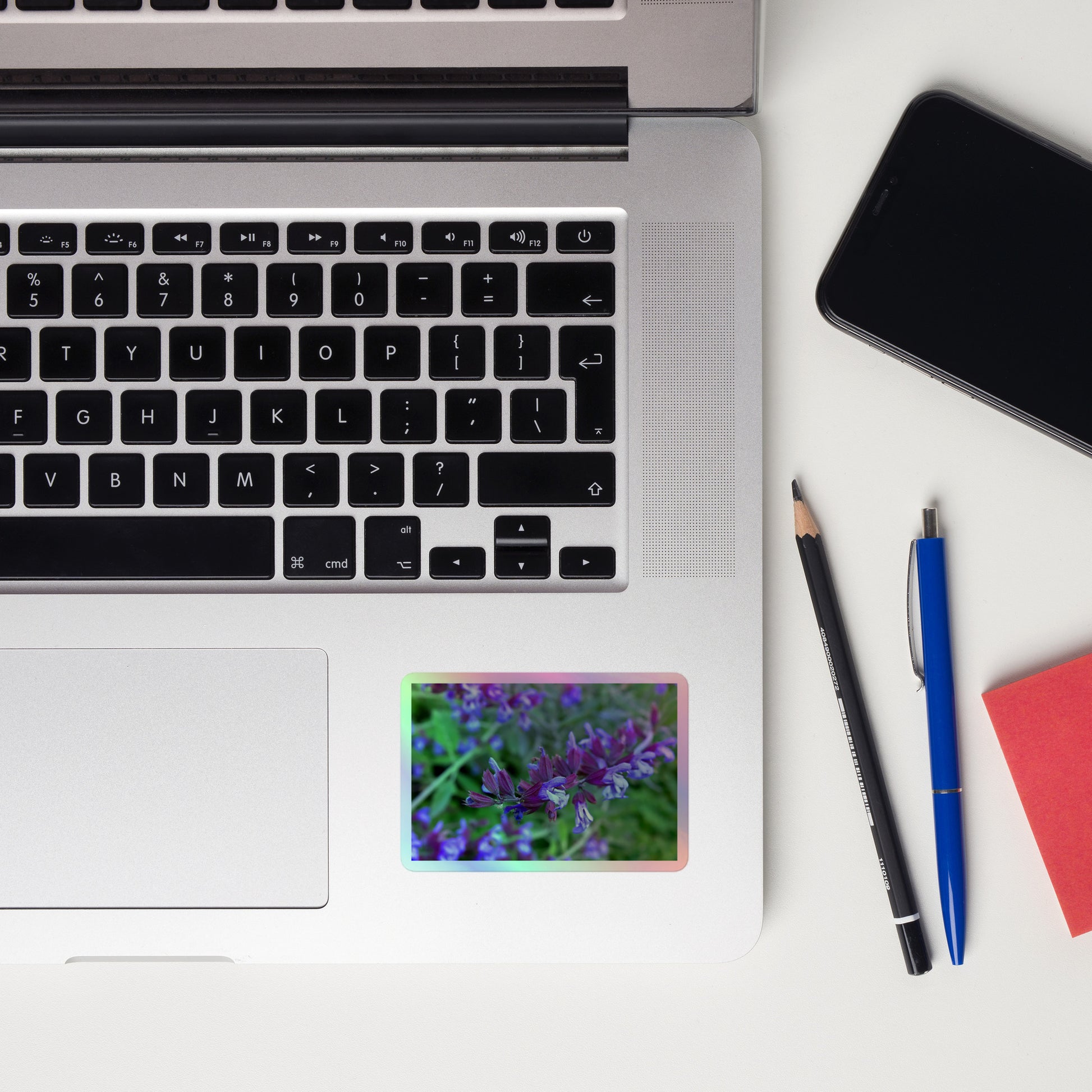 Lifestyle photo of a rectangular holographic sticker with a close-up image of bright green purple sage with green leaves, placed on a laptop to the right of the mouse pad.