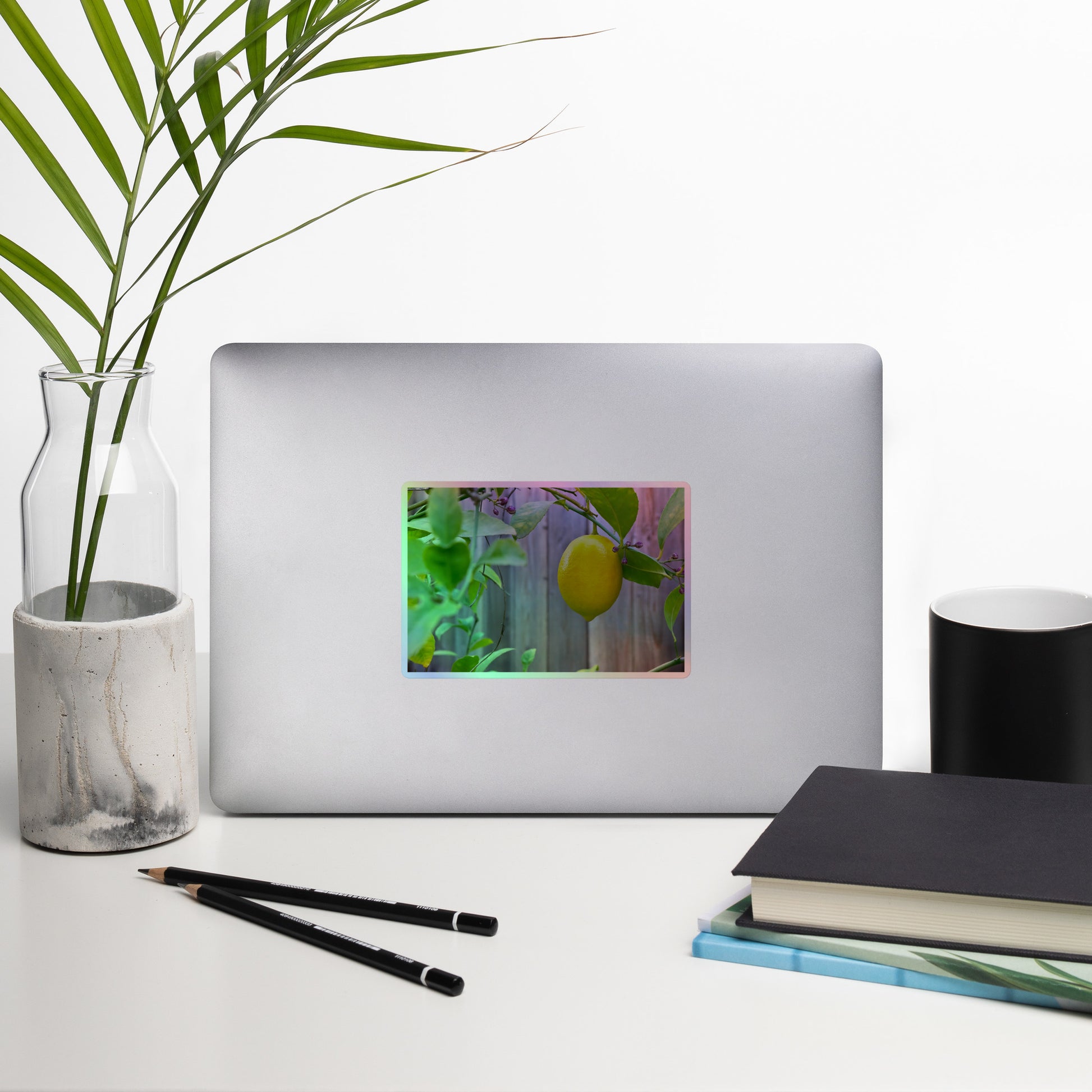Lifestyle photo of a rectangular holographic sticker with an image of a bright yellow lemon with green leaves and a wooden fence in the background, placed on a laptop with desk supplies nearby.