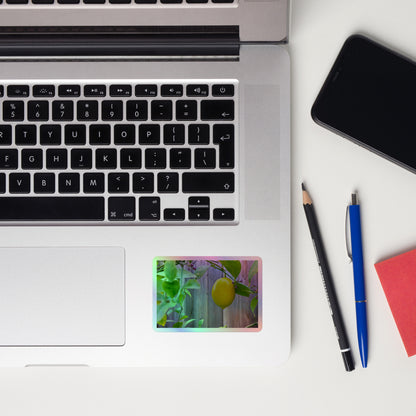 Lifestyle photo of a rectangular holographic sticker with an image of a bright yellow lemon with green leaves and a wooden fence in the background, placed on a laptop to the right of the mouse pad.