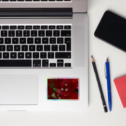 Lifestyle photo of a rectangular holographic sticker with a close-up image of a bright red bougainvillea bloom with white stamen, placed on a laptop to the right of the mouse pad.