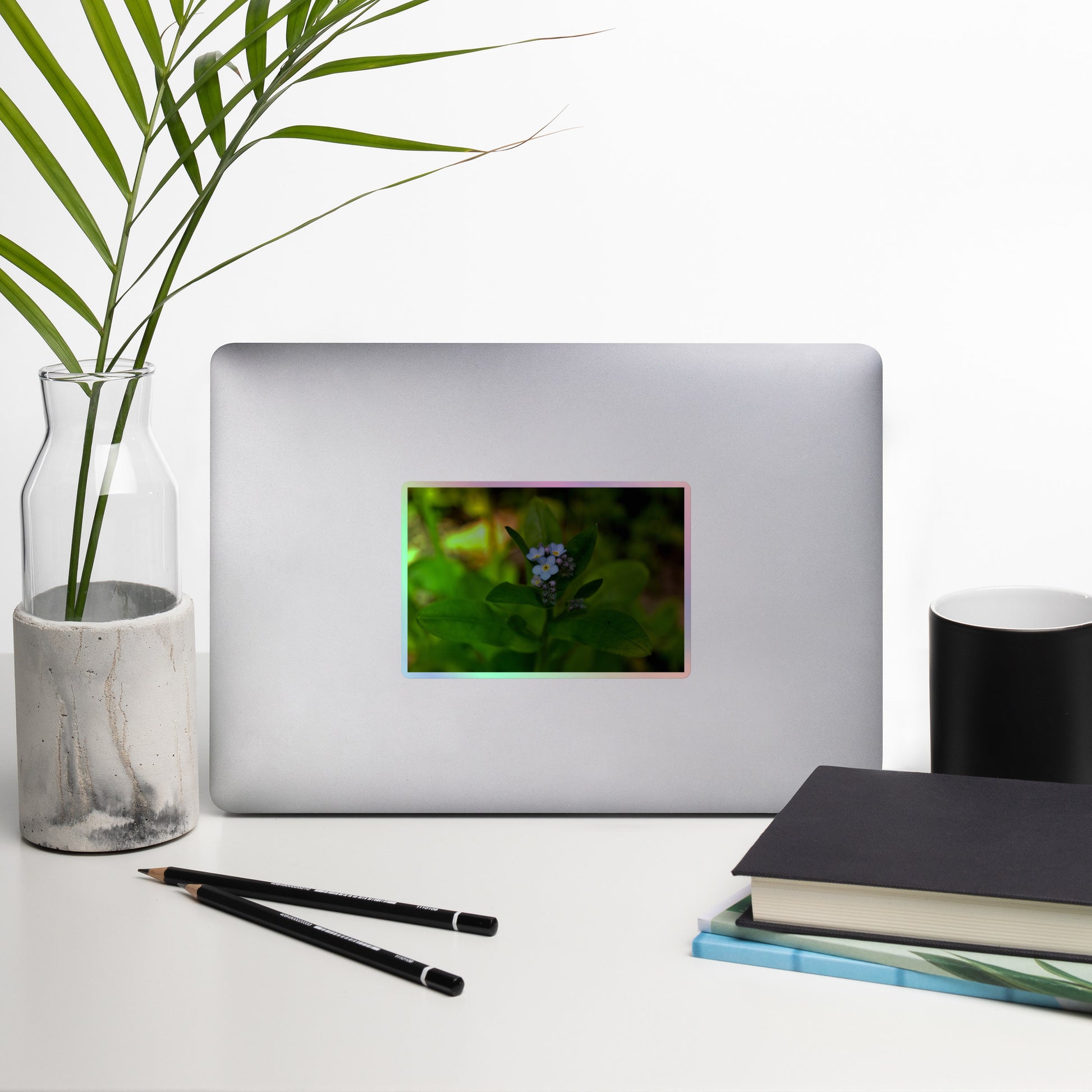 Lifestyle photo of a rectangular holographic sticker with a close-up image of a pale blue Forget-Me-Not flower in a forest background, placed on a laptop with desk supplies nearby.