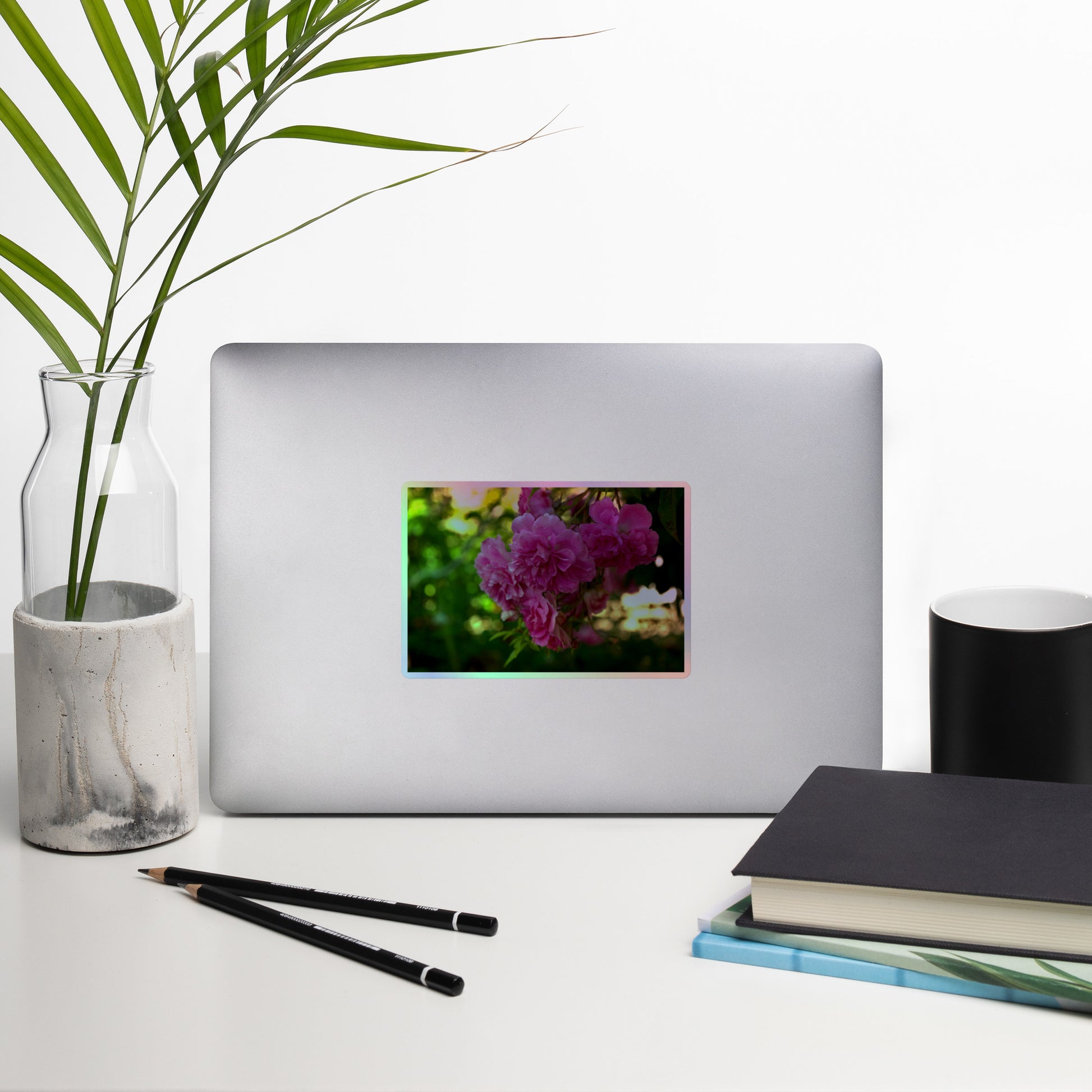 Lifestyle photo of a rectangular holographic sticker with an image of pink rose blooms in a garden, placed on a laptop with desk supplies nearby.