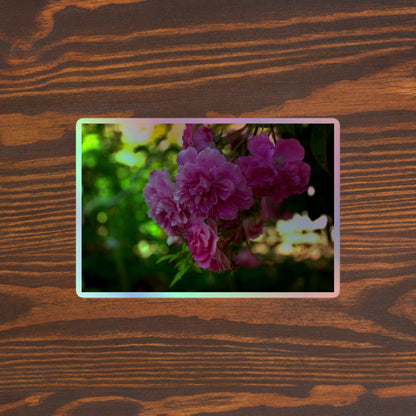 Lifestyle photo of a rectangular holographic sticker with an image of pink rose blooms in a garden, placed on a brown wooden table surface.