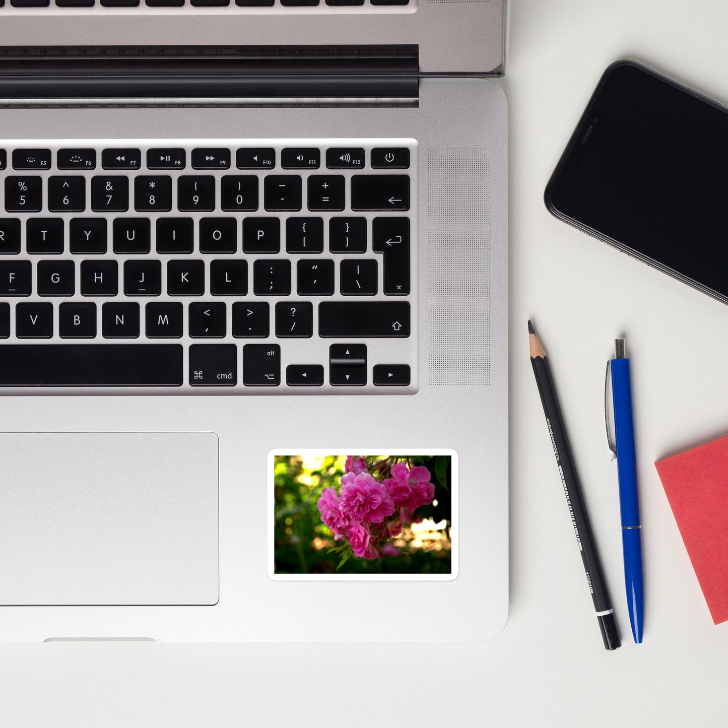 Lifestyle photo of a rectangular vinyl sticker with an image of bright pink roses in a garden, placed on a laptop to the right of the mouse pad.