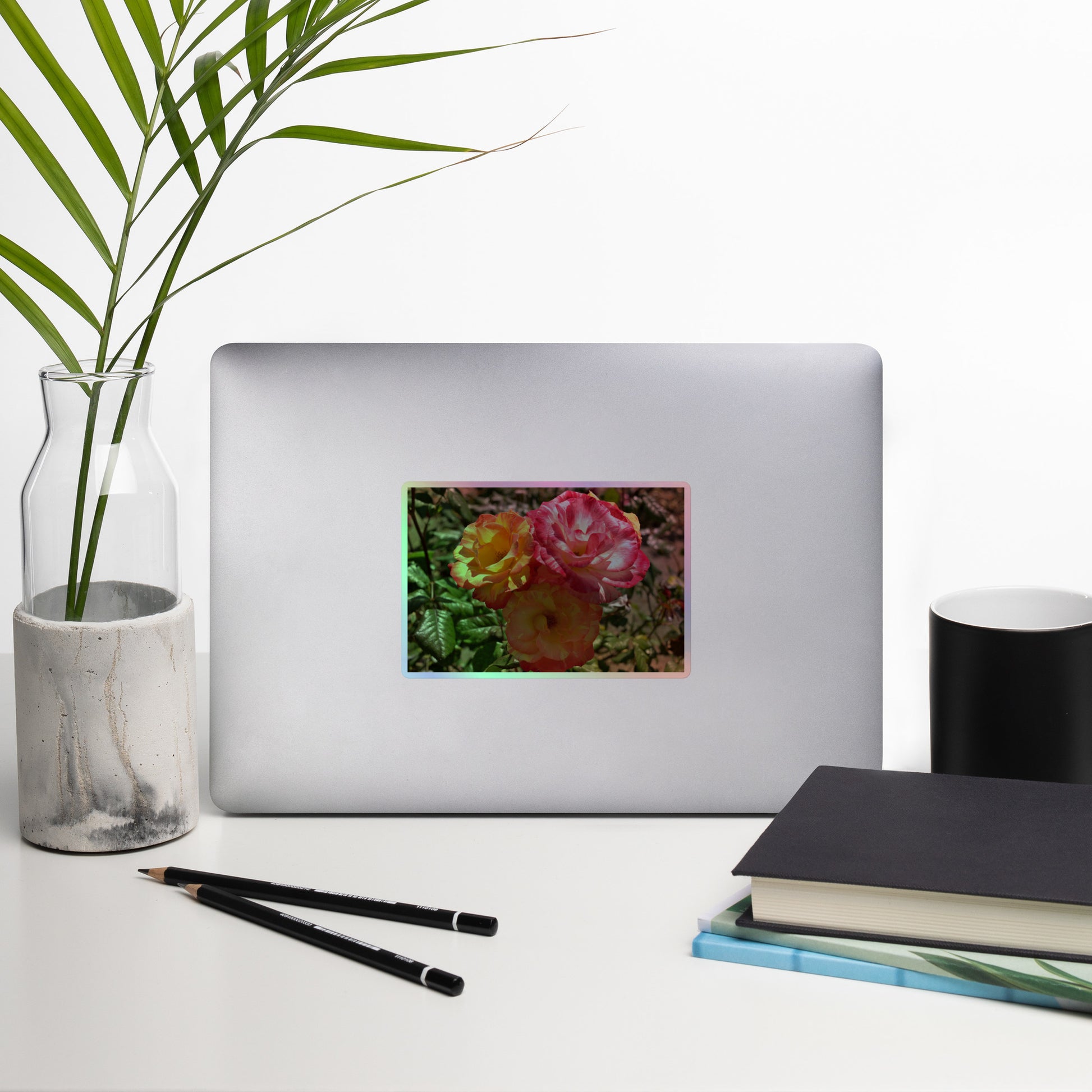 Lifestyle photo of a rectangular holographic sticker with a close-up image of three pink and yellow multicolored roses, placed on a laptop with desk supplies nearby.