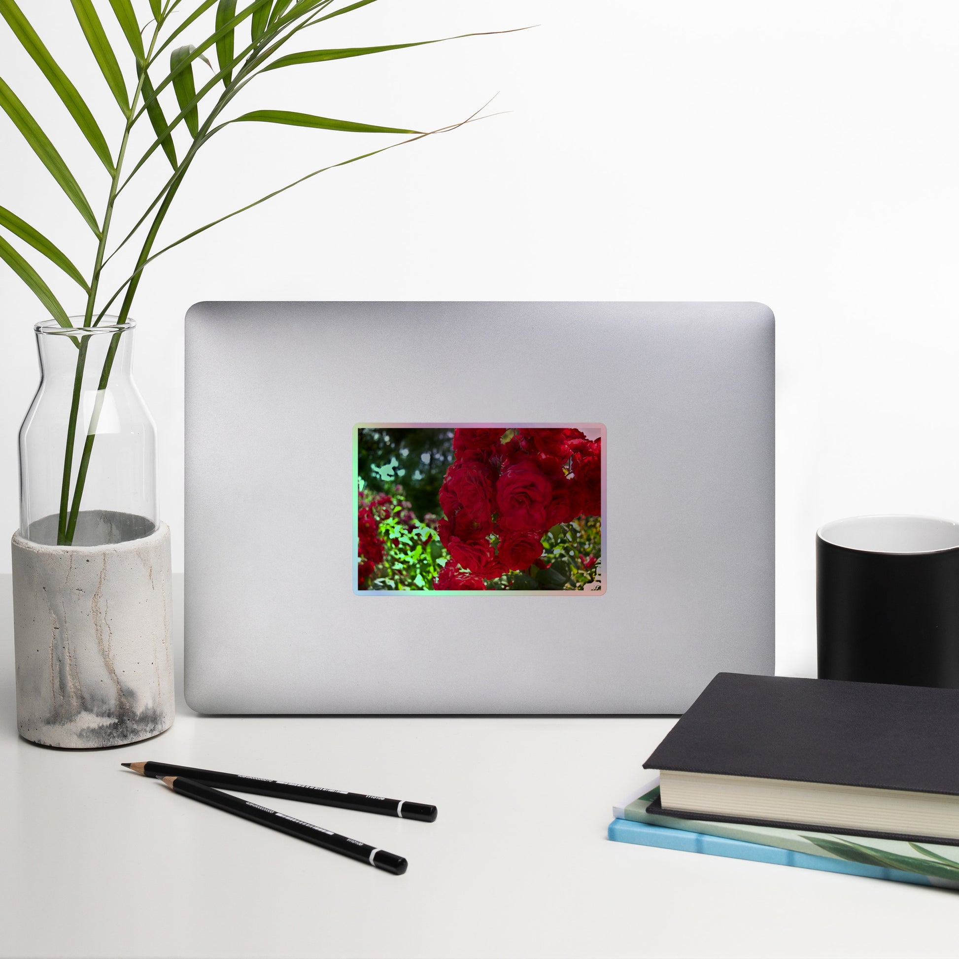 Lifestyle photo of a rectangular holographic sticker with an image of a cluster of vivid red roses, placed on a laptop with desk supplies nearby.