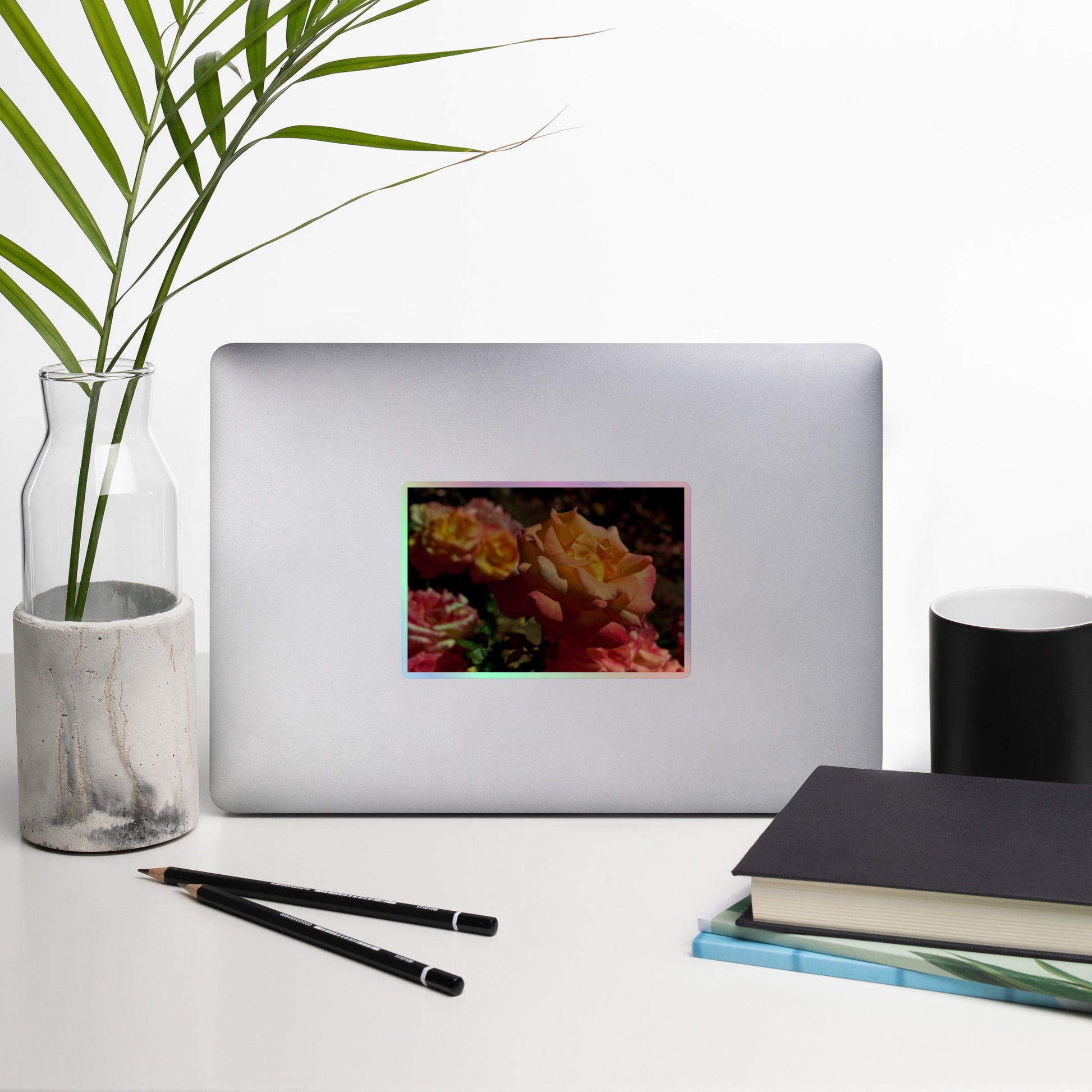 Lifestyle photo of a rectangular holographic sticker with an image of pink, orange and yellow roses in a garden, placed on a laptop with desk supplies nearby.