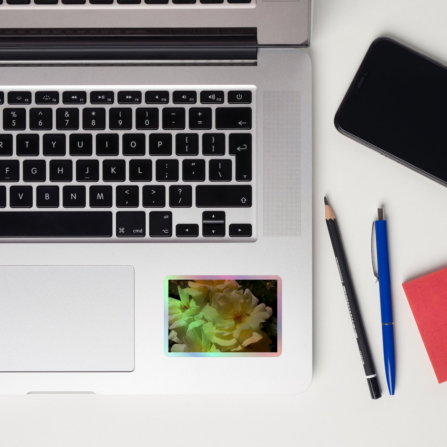 Lifestyle photo of a rectangular holographic sticker with an image of off-white rose blooms with visible stamen, placed on a laptop to the right of the mouse pad.