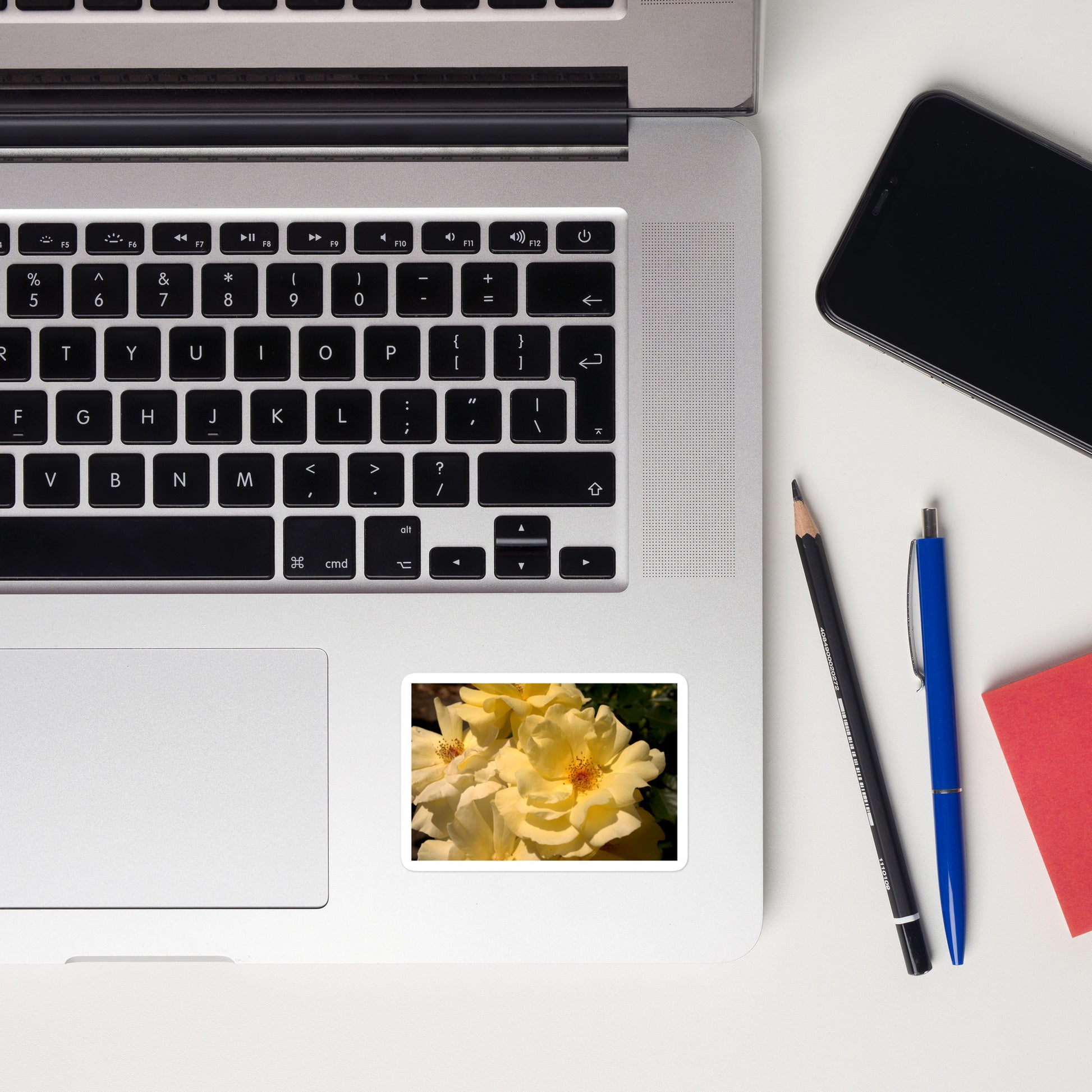 Lifestyle photo of a rectangular vinyl sticker with an image of pale yellow roses with visible stamen, placed on a laptop to the right of the mouse pad.