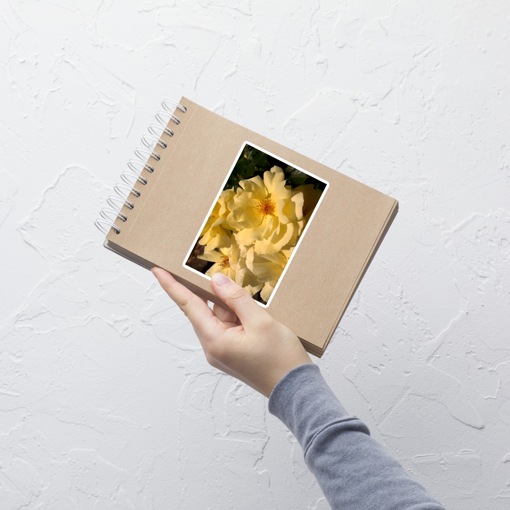 Lifestyle photo of a rectangular vinyl sticker with an image of pale yellow roses with visible stamen, placed on a small notepad.