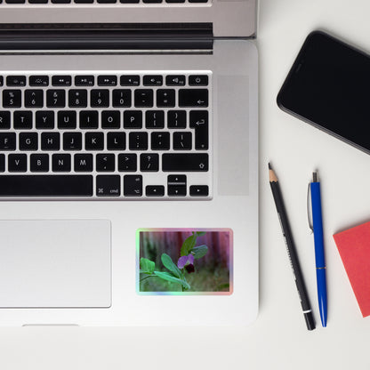 Lifestyle photo of a rectangular holographic sticker with a close-up image of a purple sweet pea with green leaves, placed on a laptop to the right of the mouse pad.