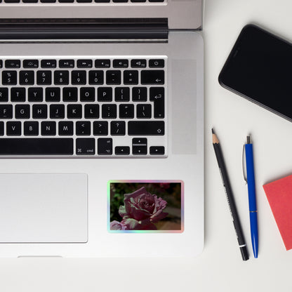 Lifestyle photo of a rectangular holographic sticker with a close up image of a light pink rose bloom, placed on a laptop to the right of the mouse pad.