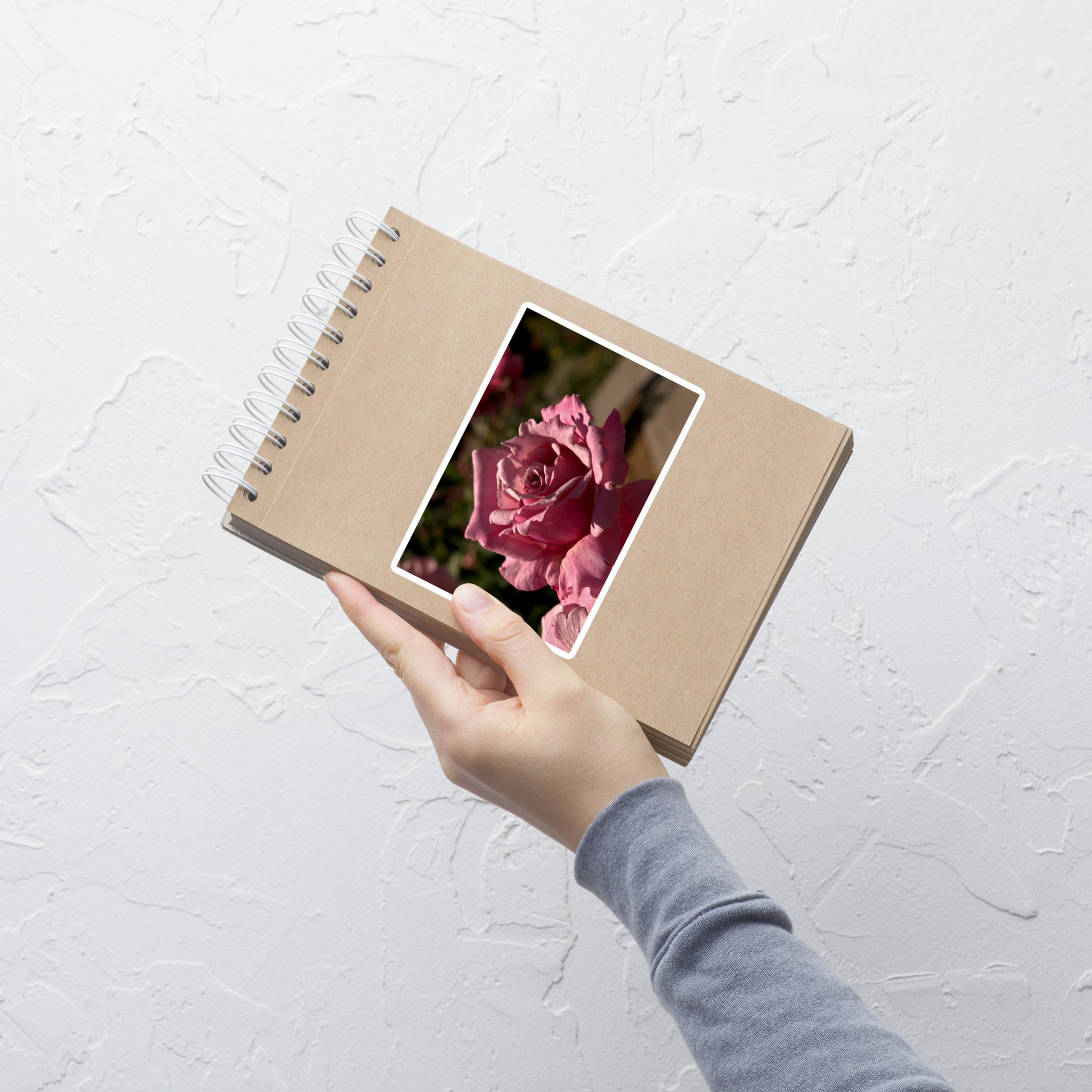 Lifestyle photo of a rectangular vinyl sticker with a close-up image of a bright pink rose bloom, placed on a small notepad.