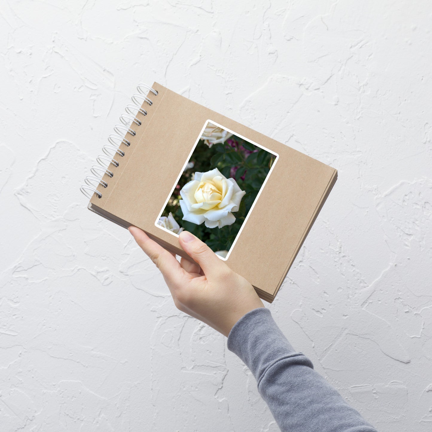 Lifestyle photo of a rectangular vinyl sticker with a close-up image of a white rose bloom with a pale yellow center, placed on a small notepad.