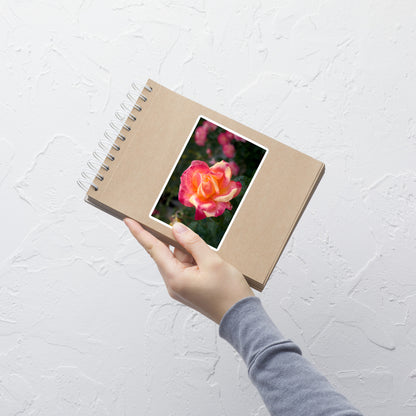 Lifestyle photo of a rectangular vinyl sticker with an image of a pink and yellow rose bloom with dark green leaves, placed on a small notepad.