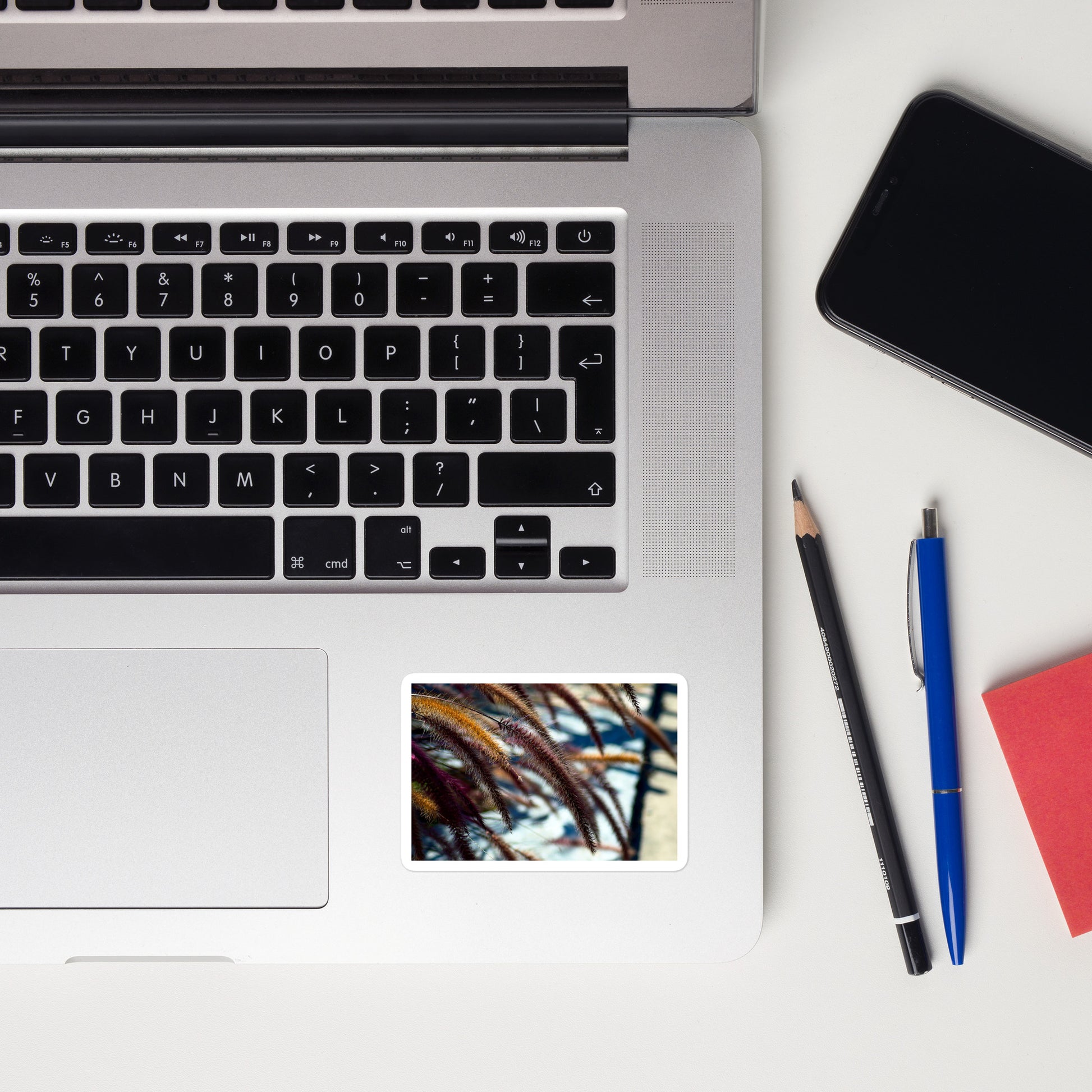 Lifestyle photo of a rectangular vinyl sticker with an image of yellow and purple foxtails, placed on a laptop to the right of the mouse pad.