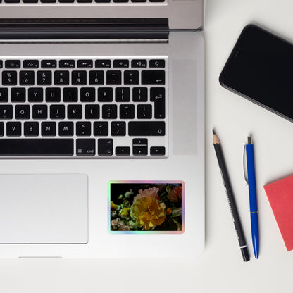 Lifestyle photo of a rectangular holographic sticker with an image of pale yellow and pink rose blooms in a garden, placed on a laptop to the right of the mouse pad.