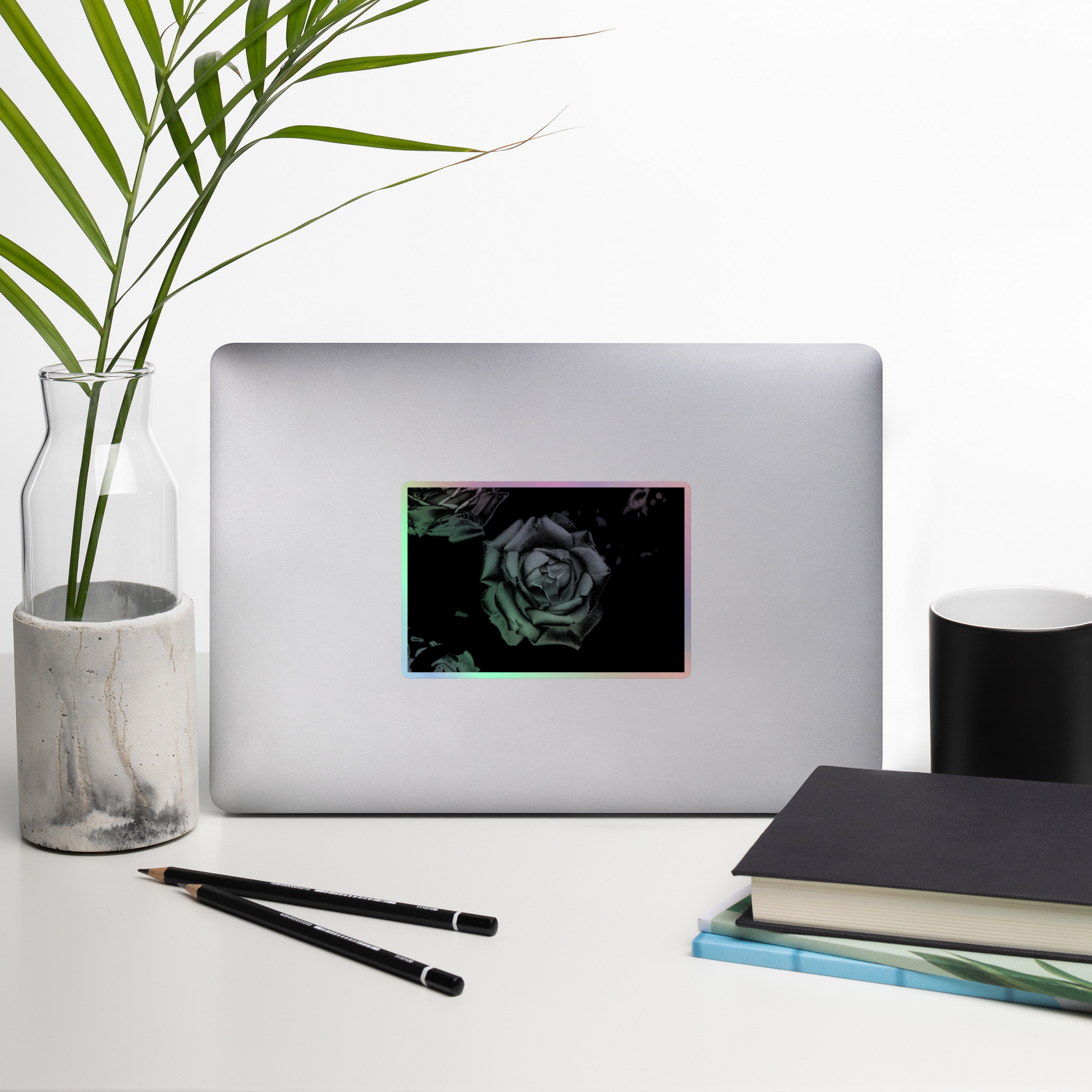 Lifestyle photo of a rectangular holographic sticker with an image of a grey charcoal rose design, placed on a laptop with desk supplies nearby.