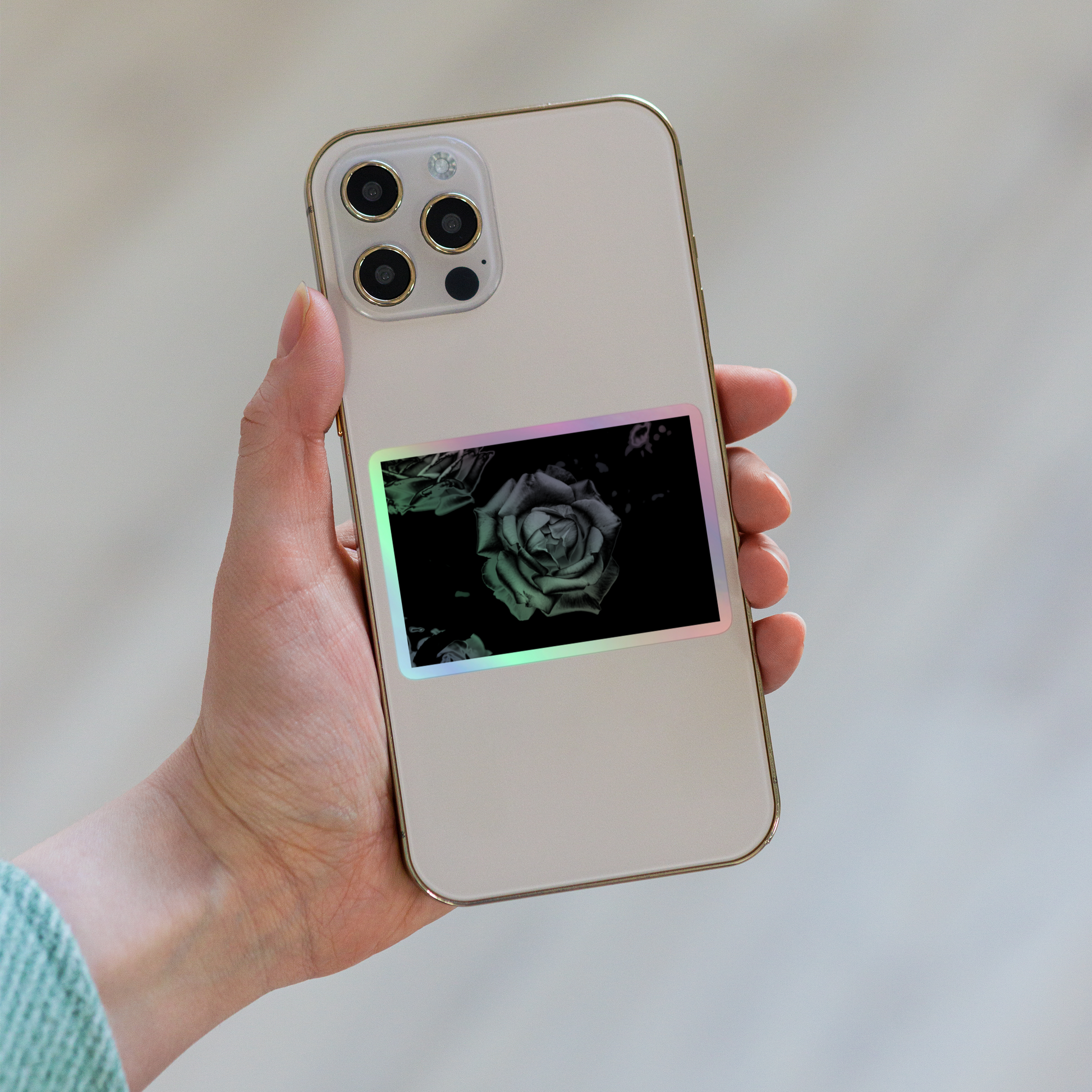 Lifestyle photo of a rectangular holographic sticker with an image of a grey charcoal rose design, placed on the back of a phone.