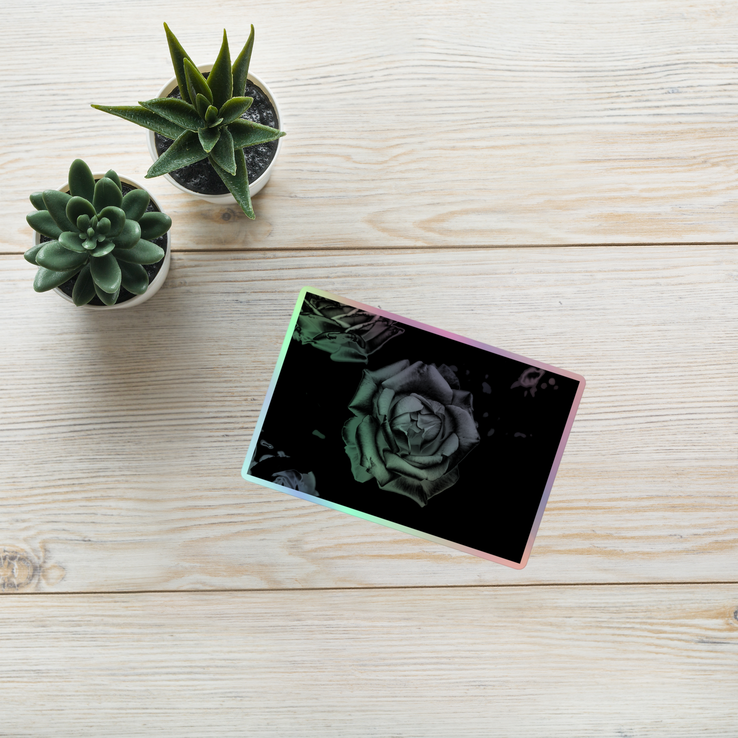 Lifestyle photo of a rectangular holographic sticker with an image of a grey charcoal rose design, placed on a table with two small succulents.