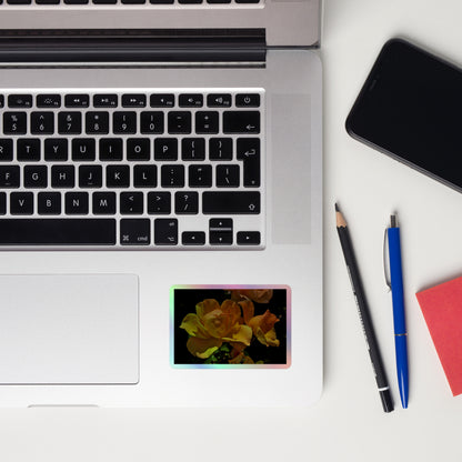 Lifestyle photo of a rectangular holographic sticker with a high-contrast image of vivid yellow roses against a dark background, placed on a laptop to the right of the mouse pad.