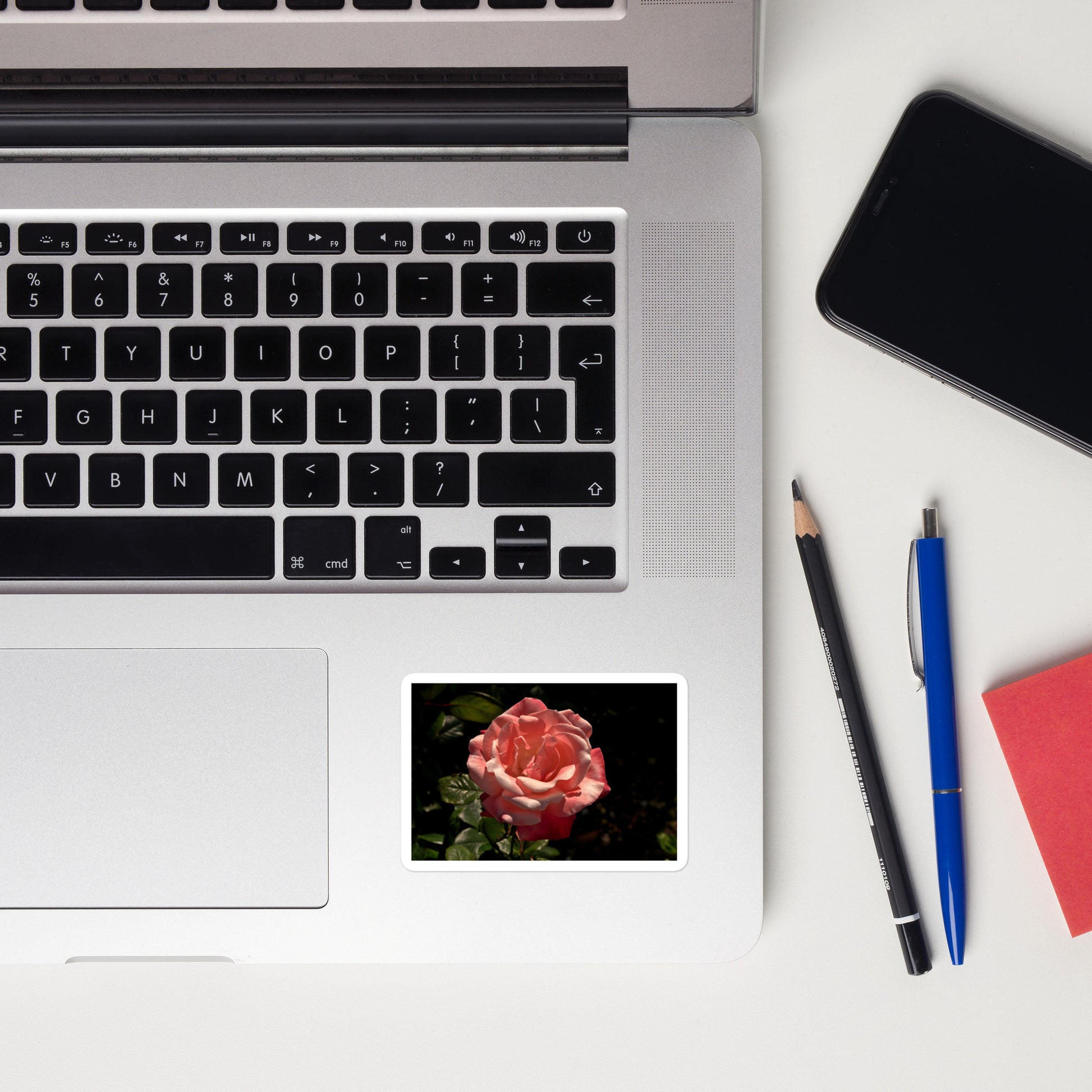 Lifestyle photo of a rectangular vinyl sticker with a high contrast image of a light pink rose bloom with dark green leaves, placed on a laptop to the right of the mouse pad.